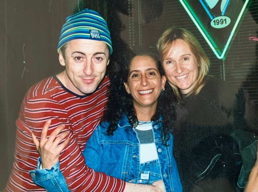 Three friends smiling and posing together indoors, with neon signs in the background.