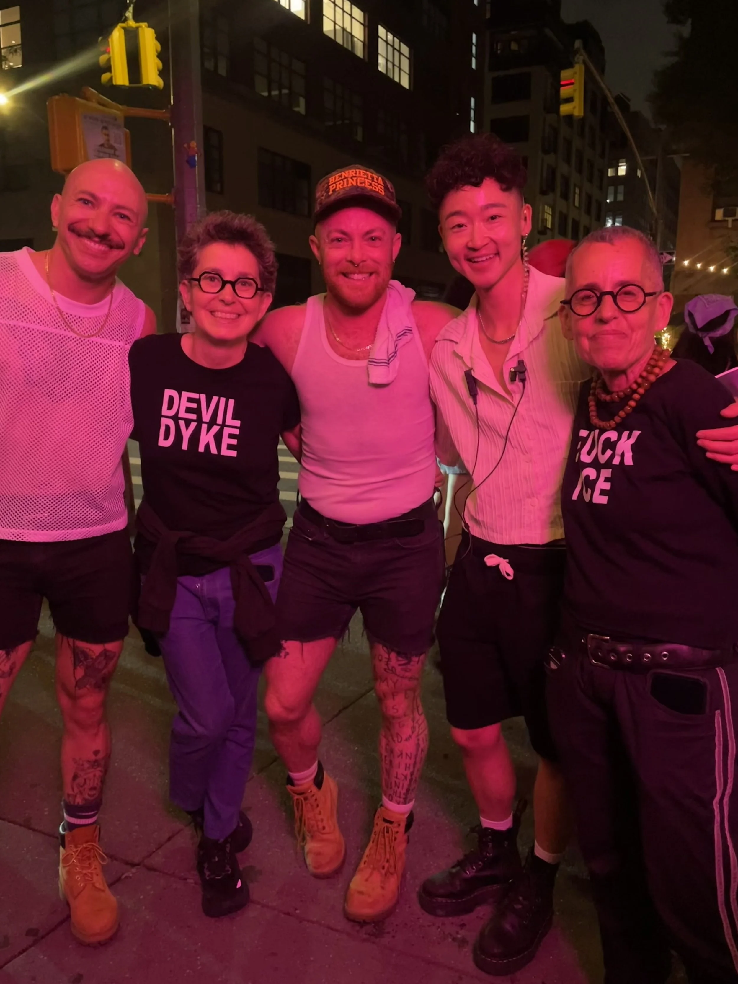 Group of five diverse people standing together on a city street at night, smiling at the camera, wearing colorful and eclectic clothing, illuminated by pink and red lighting.