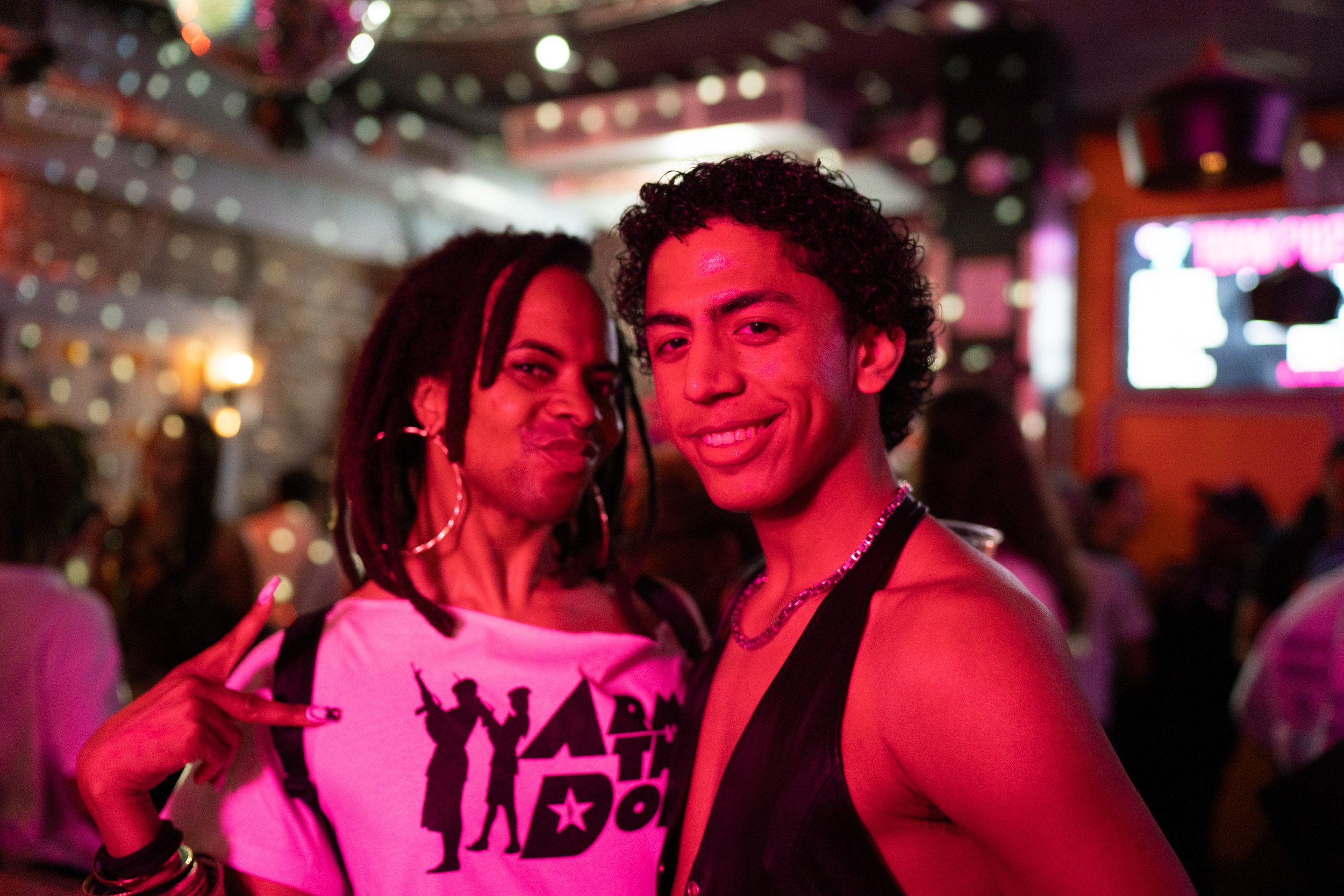 Two people at a lively indoor party or club, with colorful, blurred lights and a neon sign in the background. They are standing close together, smiling, with one person showing a peace sign. The scene has a fun, vibrant atmosphere.