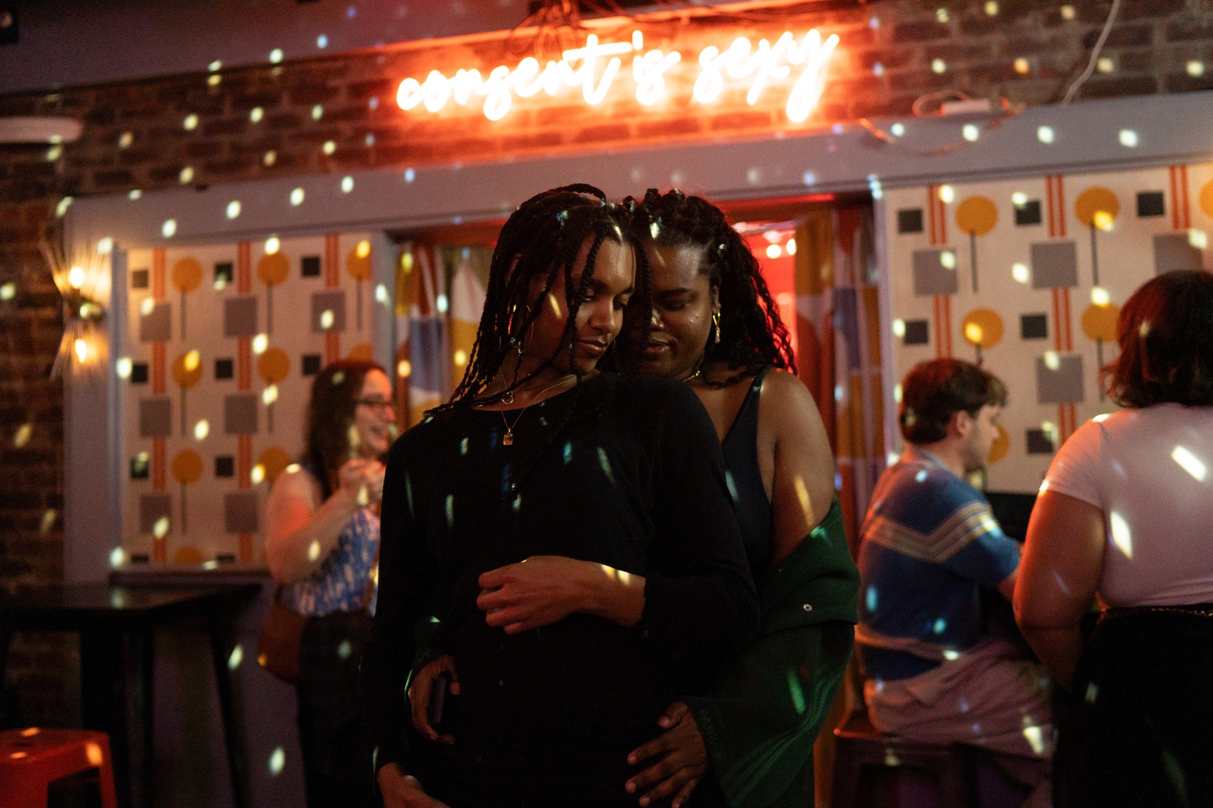 Two women dancing closely in a dimly lit room with colorful lights and a neon sign that reads 'confetti is sexy' in the background.