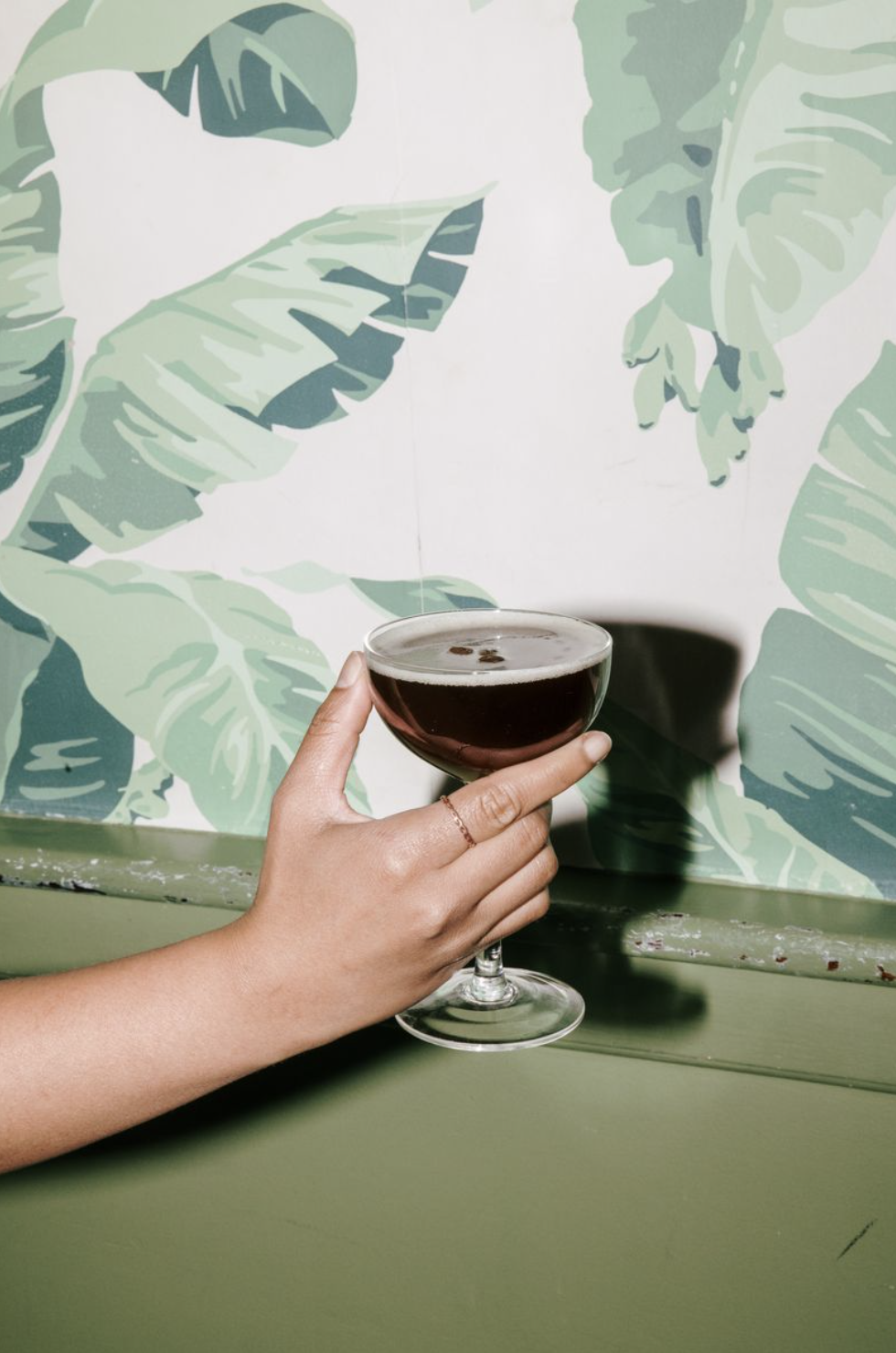 Hand holding a cocktail glass with a dark drink, against a green leafy mural background.