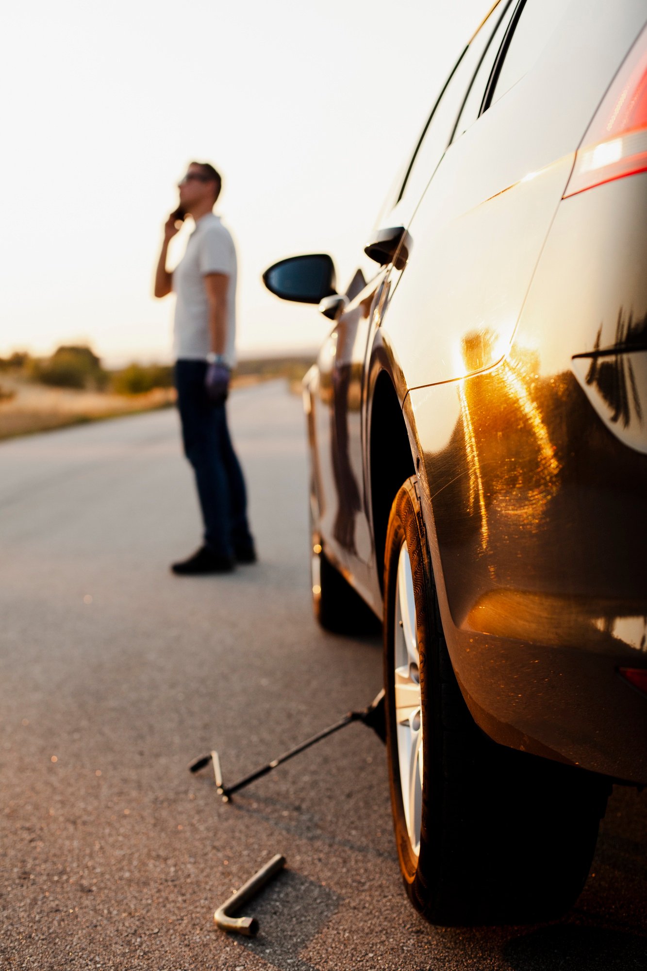 A man standing next to a car that is broken down on the side of the road during sunset, with a tire jack and tools on the ground.