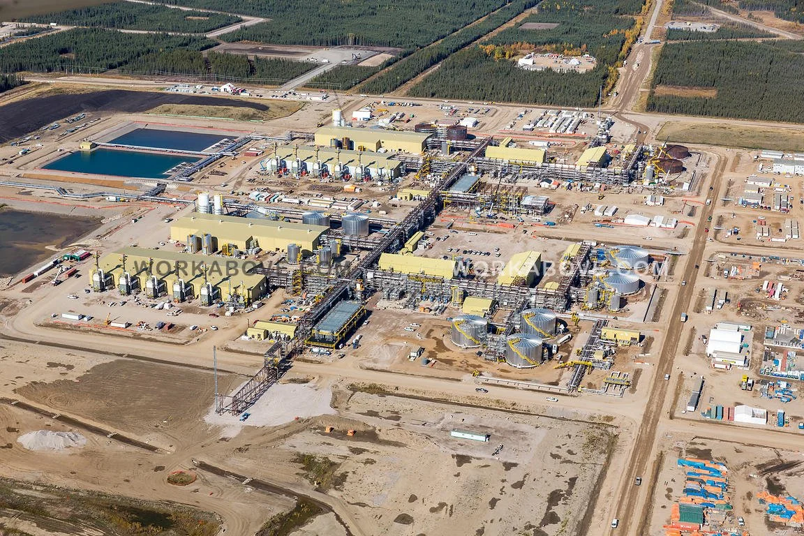 Aerial view of an industrial plant with multiple large tanks, pipelines, and buildings in a rural area surrounded by forests and dirt roads.
