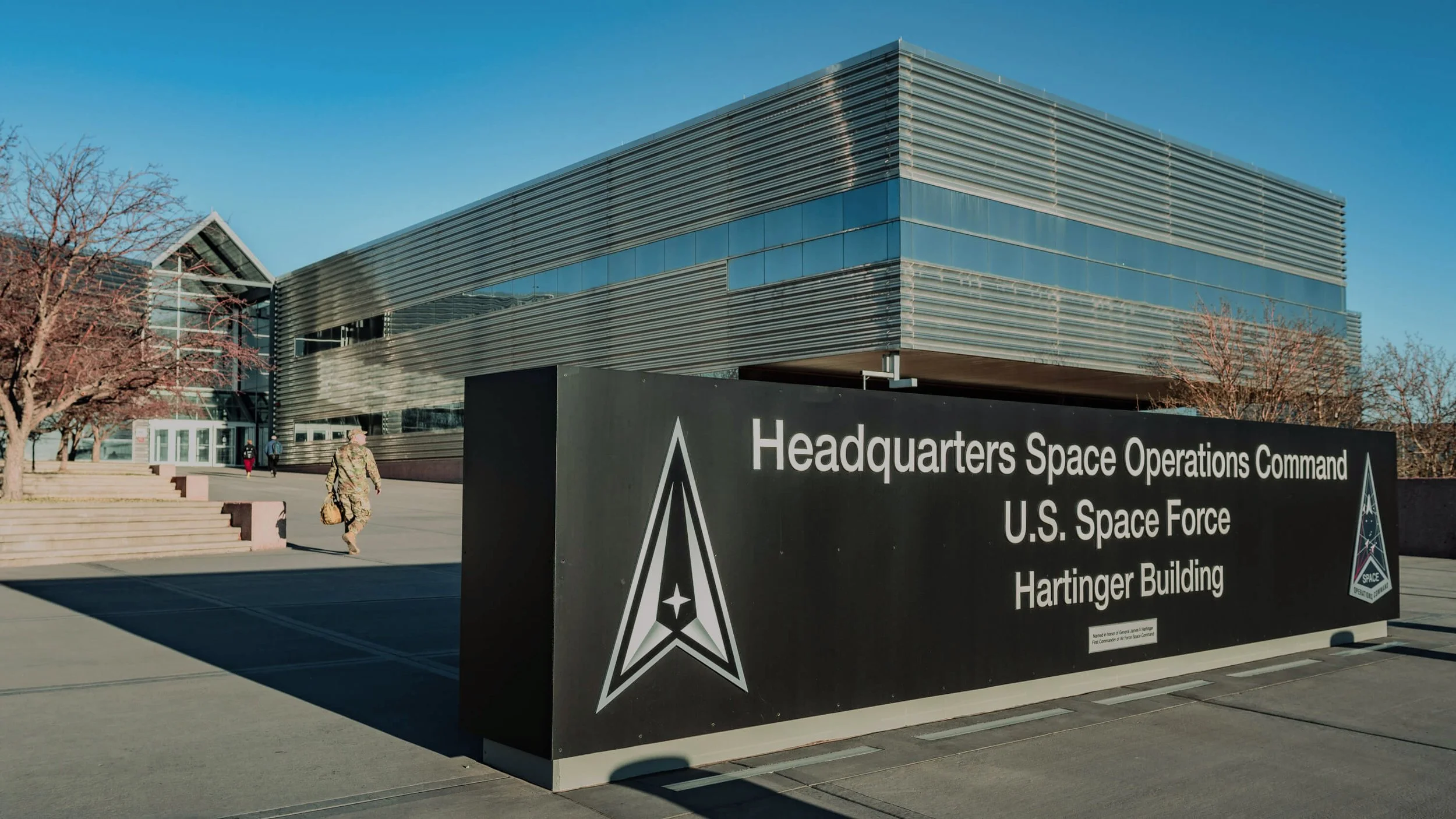 The Hartinger Building, U.S. Space Force headquarters, with a black sign in front displaying the organization name and logo, and a modern glass and metal building in the background under a clear blue sky.