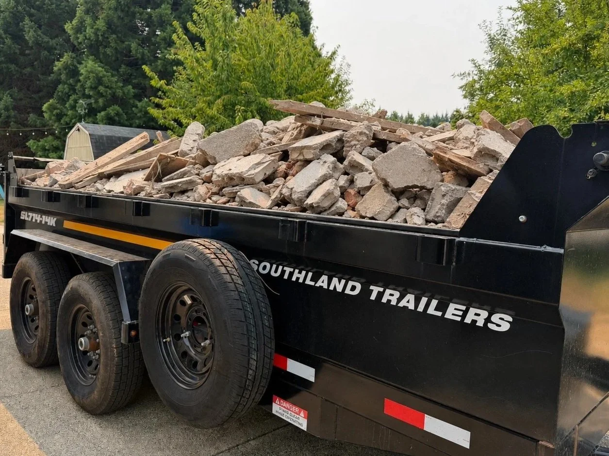 A black trailer labeled 'Southland Trailers' filled with broken concrete and wooden debris, parked on a paved surface with green trees in the background.