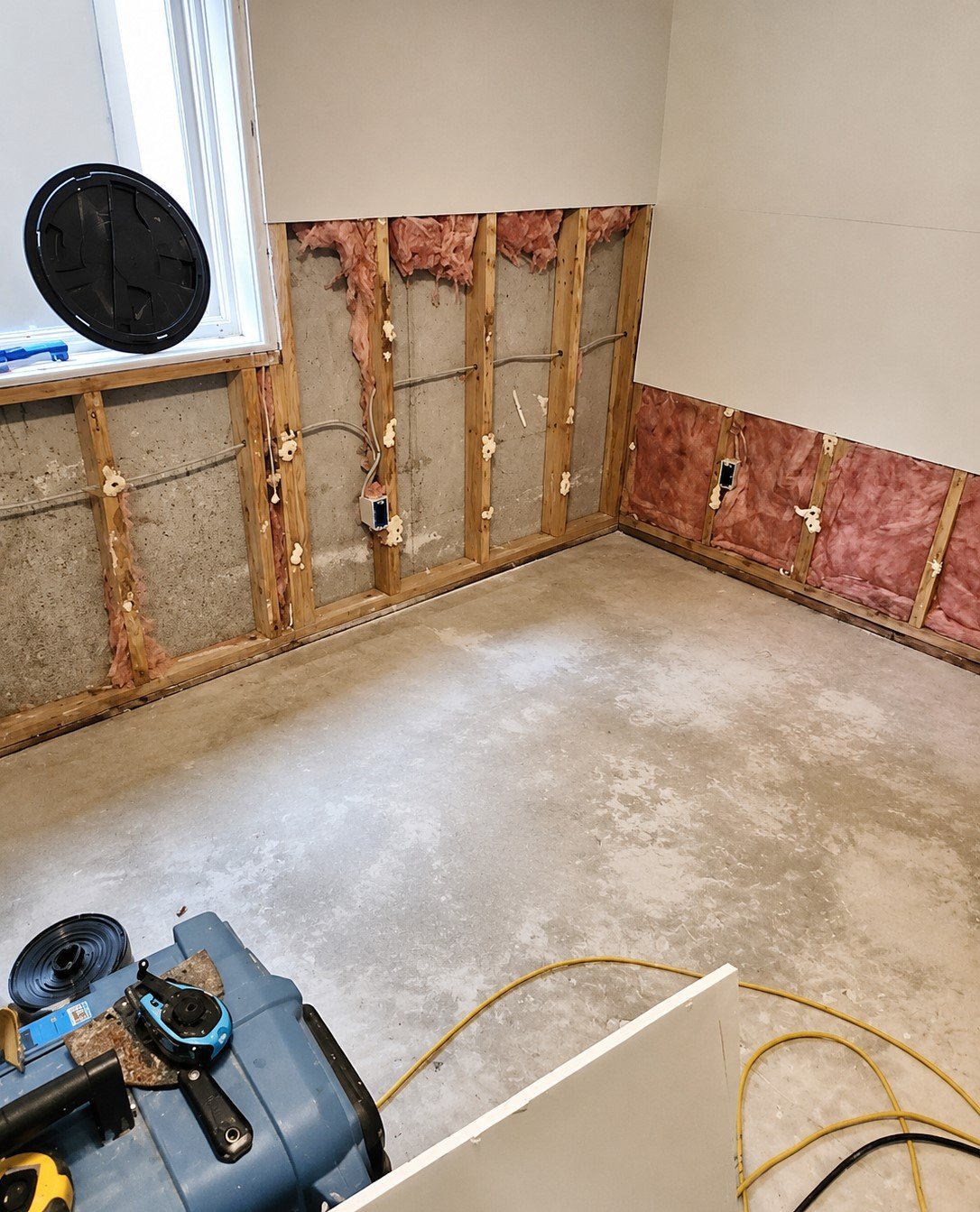 A room under renovation with exposed wall studs, insulation, and electrical wiring. The floor is unfinished concrete, and construction tools are in the workspace.