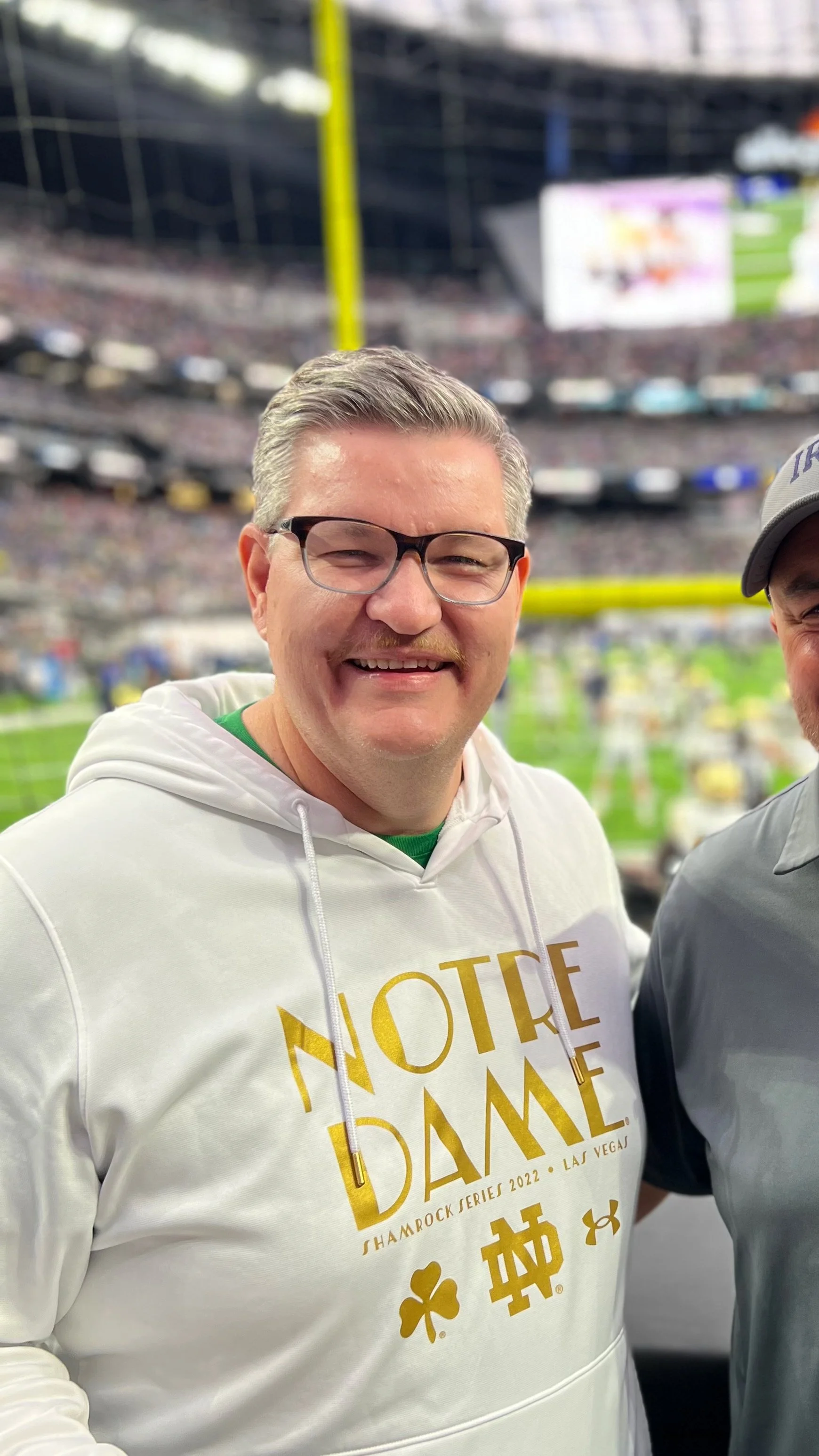 A smiling man with glasses and a mustache wearing a white hoodie with gold text, at a sports stadium during a game.