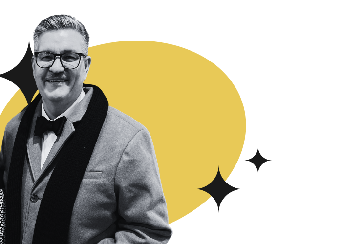 A man in a tuxedo with a bow tie, wearing glasses, smiling, standing against a yellow background with some green and black design elements.