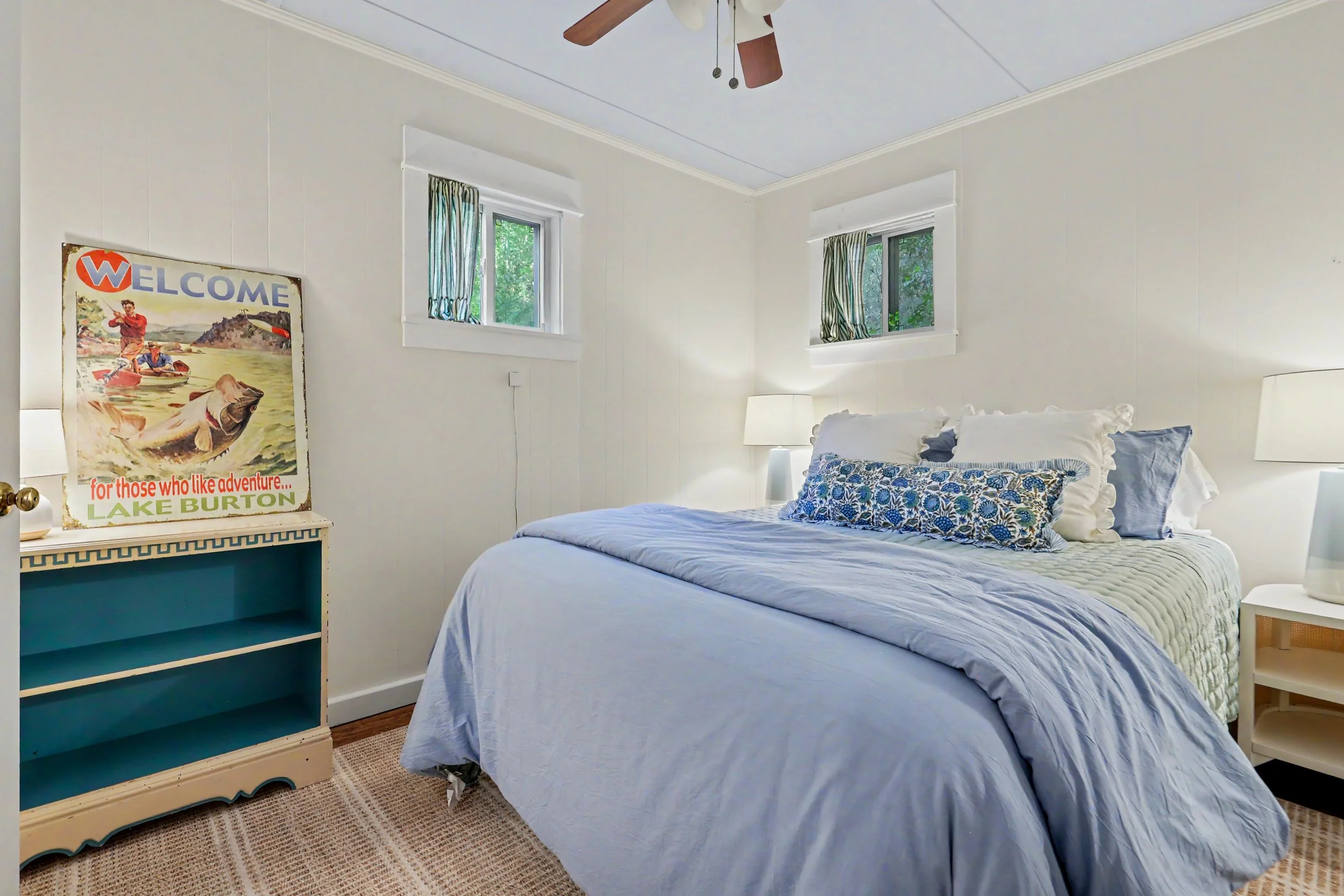 A bedroom with a white bed covered in a light blue quilt and decorative pillows. There are two white nightstands with lamps, and a vintage-style poster featuring a fishing scene on a white piece of furniture. The room has two small windows with patte