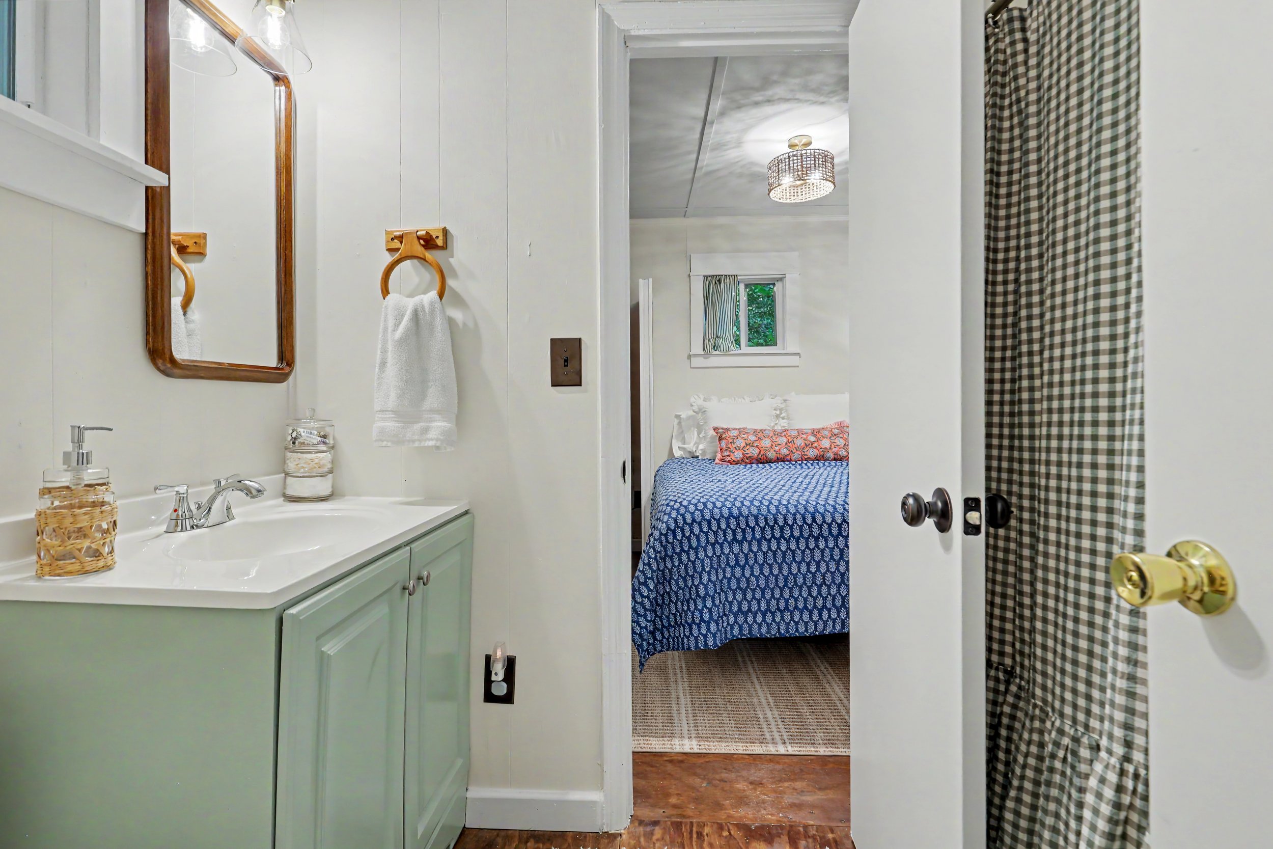 A bathroom view looking into a bedroom through an open door. The bathroom has light green cabinets, a white sink, and decorative items on the countertop. The bedroom has a bed with a blue and white patterned bedspread, colorful pillows, a window with