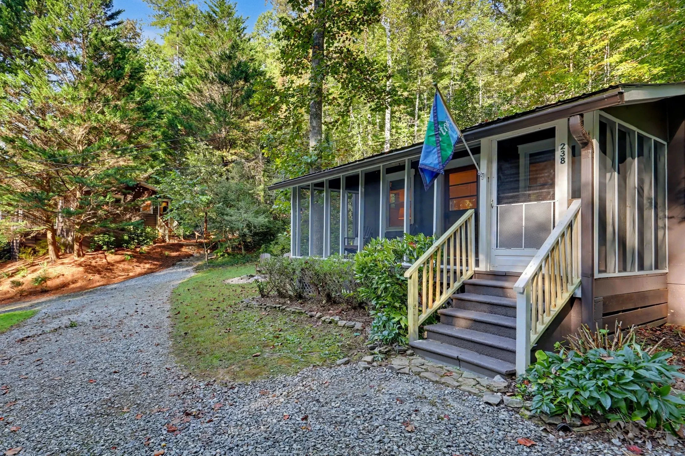 A view of a Lake Burton cabin rental house with a welcoming porch, surrounded by a lush wooded area with tall trees. A gravel pathway leads up to the front steps, and a flag is flying on the porch.