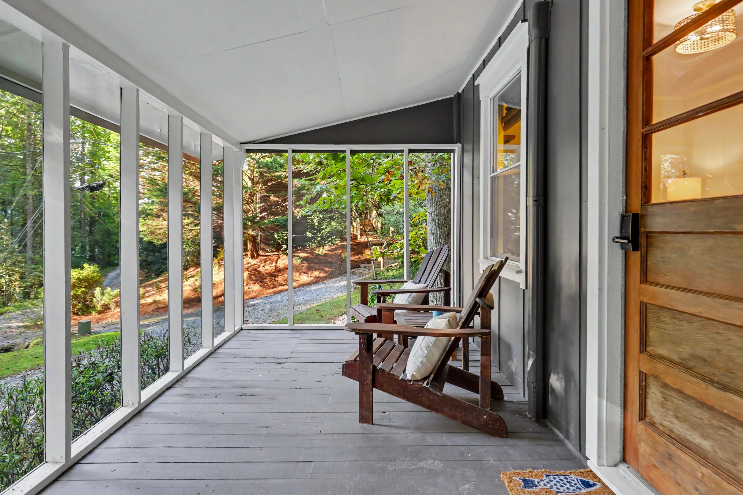 View of a covered porch with wooden chairs and a door, overlooking a lush green yard with trees.