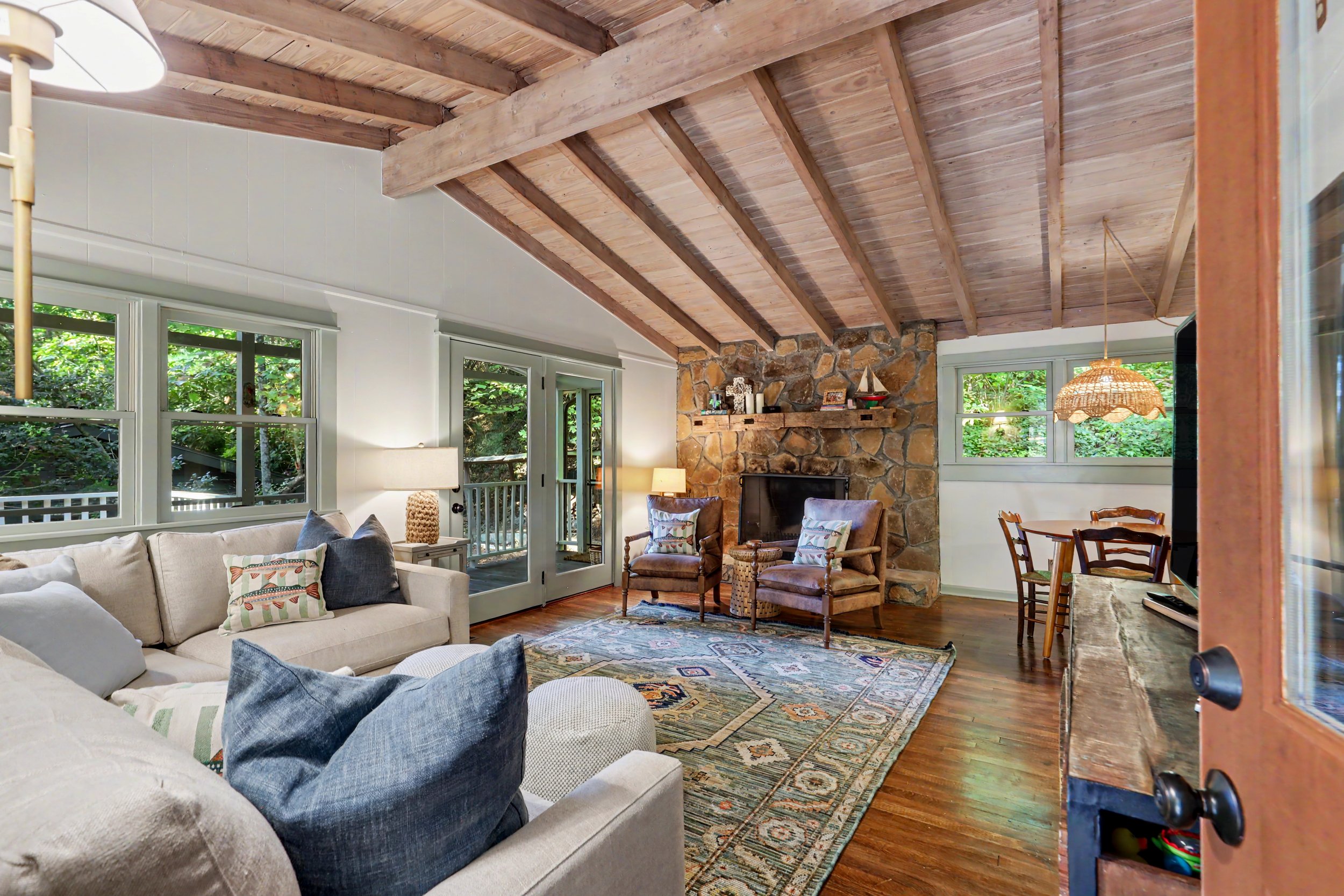 North Georgia mountain cabin rental living room with beige sofa, wooden ceiling, stone fireplace, large windows showing greenery outside, wooden chairs, and a colorful rug.