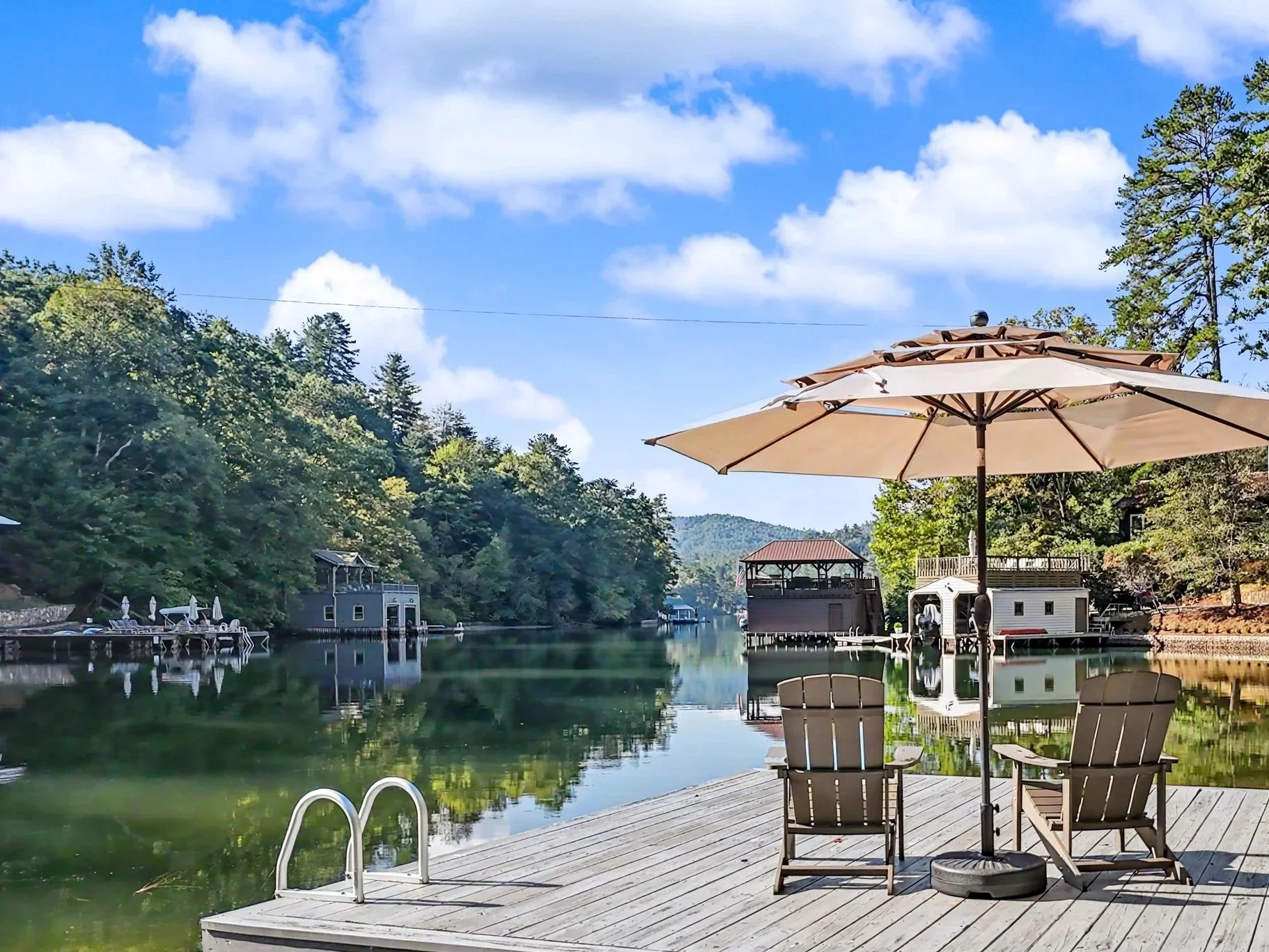 A Lake Burton view with a wooden dock and two Adirondack chairs under a large beige patio umbrella. The calm water reflects trees, houses, and a sky with scattered clouds on a sunny day.