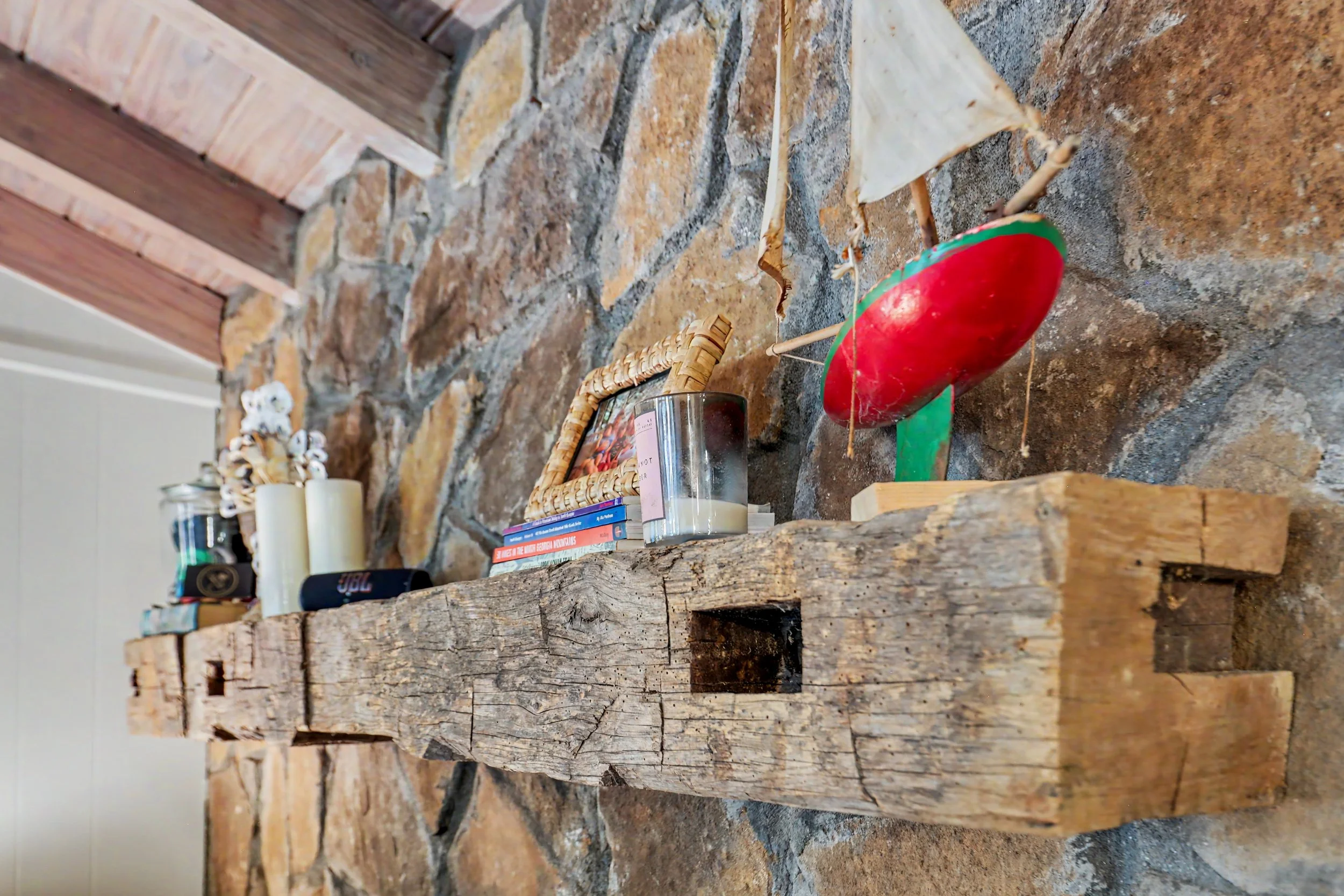 Close-up of a rustic wooden mantle with candles, books, and a small photograph frame placed on it. The background includes a stone wall.