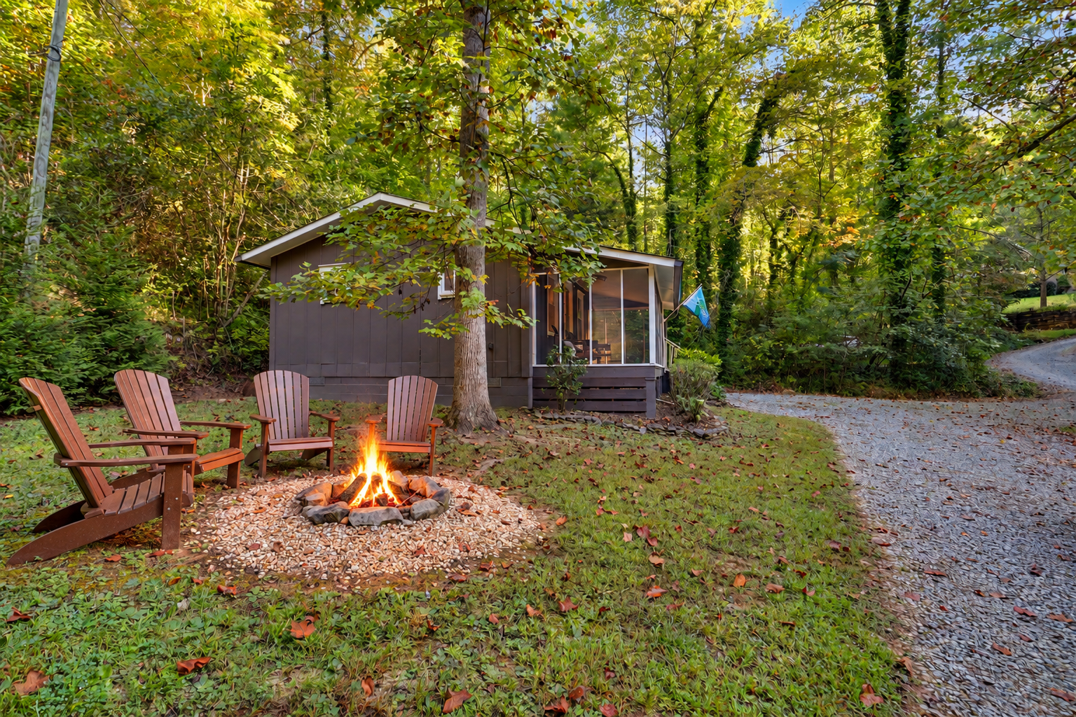 A Lake Burton mountain cabin backyard scene with four wooden Adirondack chairs around a campfire in a rocky fire pit, in front of a small gray house with a screened porch, surrounded by green trees and a winding gravel path.