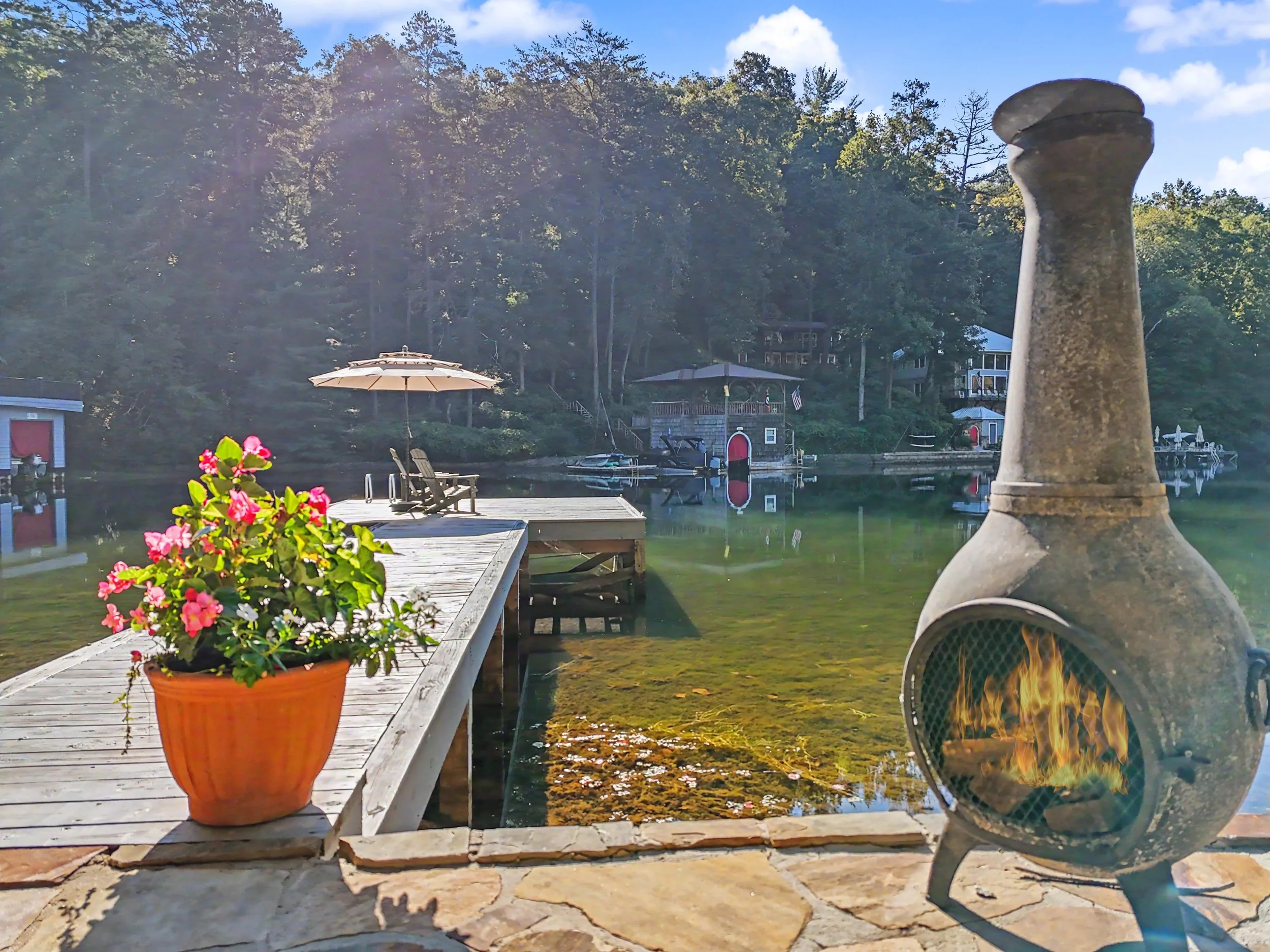 A lakeside scene with a wooden dock, pink flowers in a terracotta pot, a outdoor umbrella, a stone patio, a decorative stone column, and a fire pit with flames, with houses and trees in the background under a partly cloudy sky.