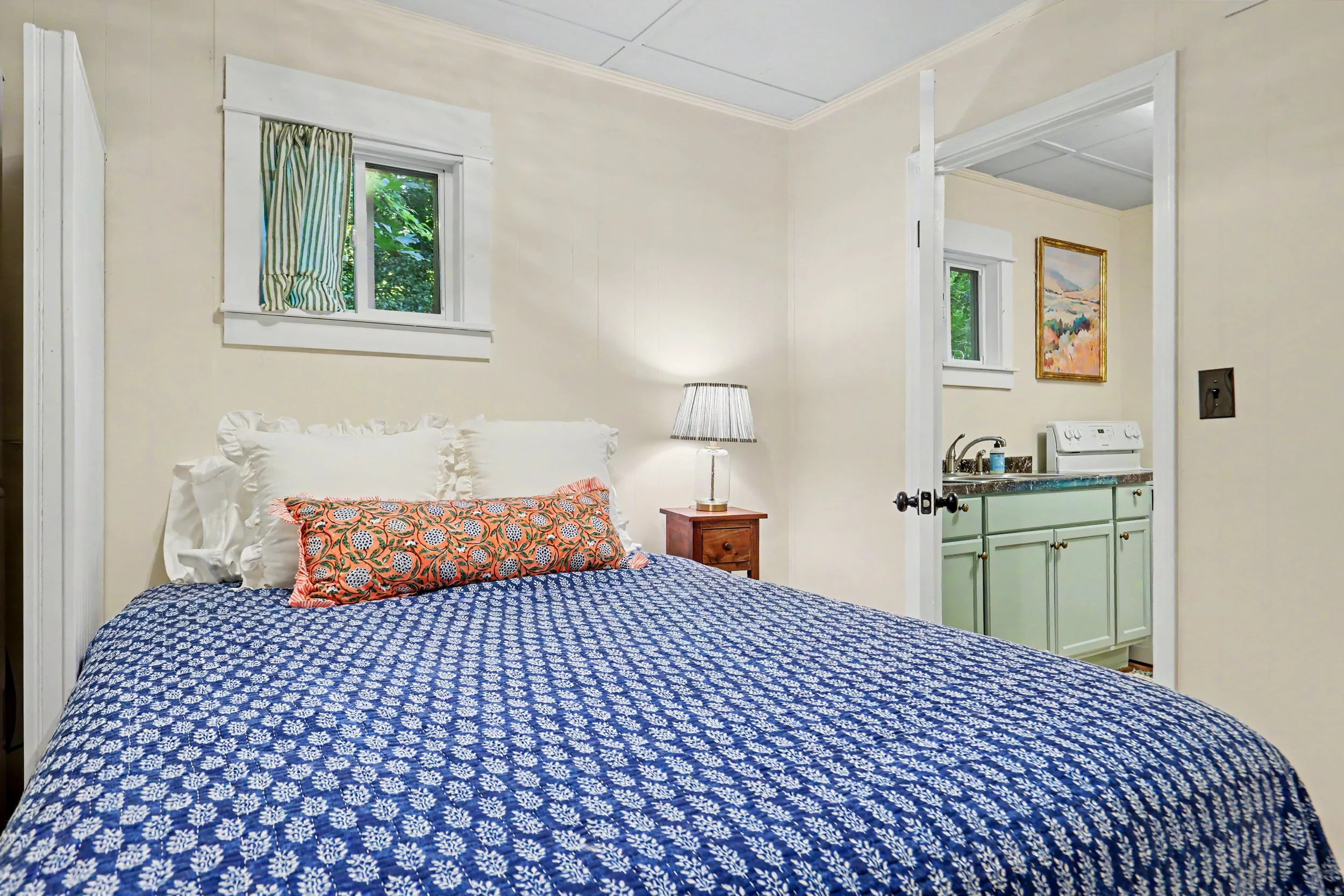 A cozy bedroom with a bed covered in blue and white patterned bedding and stacked pillows, a small wooden nightstand with a lamp, and an adjacent small kitchen area with green cabinets and a stove, visible through an open doorway.