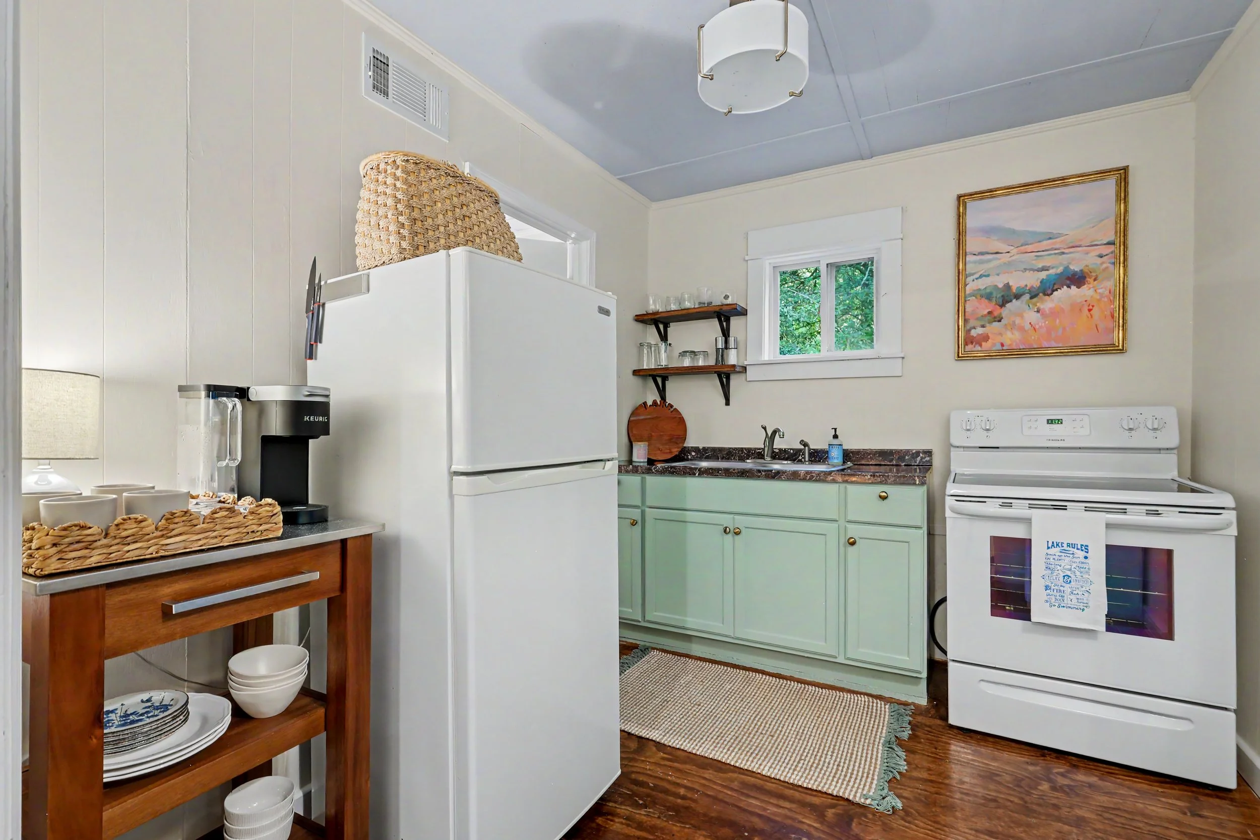 Small kitchen with mint green cabinets, white stove, white refrigerator, wooden shelves with glasses, colorful framed painting, window with trees outside, woven basket on top of refrigerator, Keurig coffee maker, and linen towels hanging from oven ha