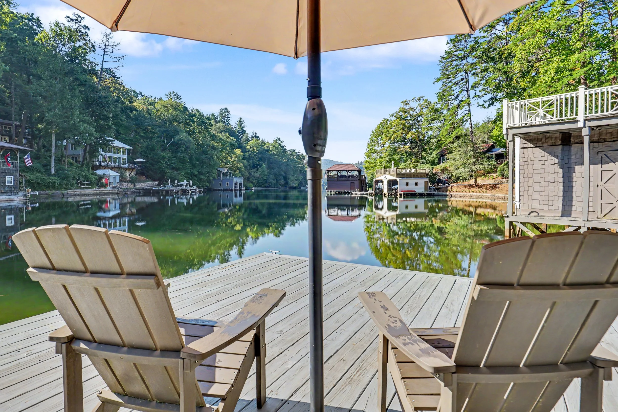 View from a lakeside dock on Perrin Cove with Adirondack chairs and a large umbrella, overlooking a calm lake with trees and boathouses.
