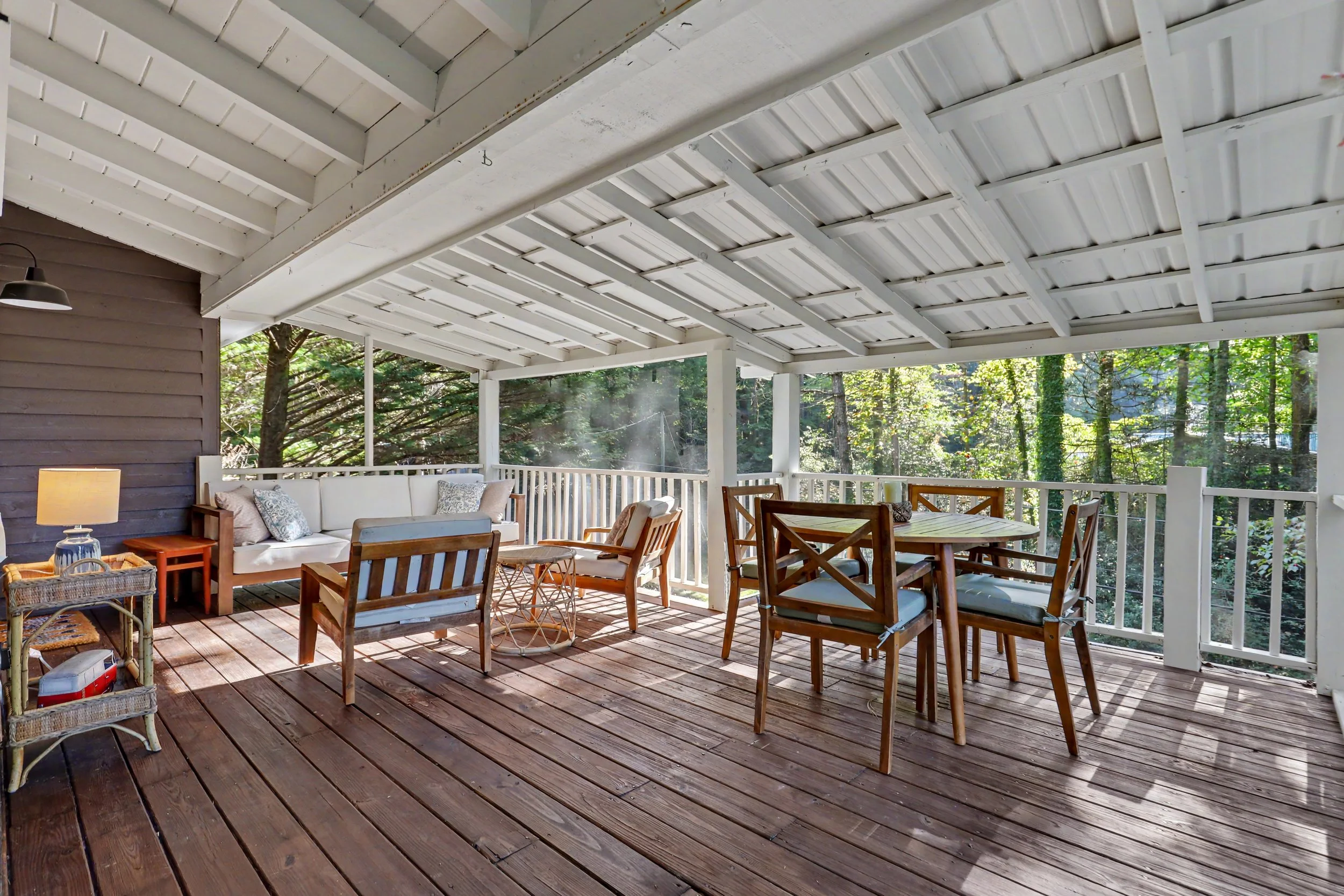 Covered porch with wooden flooring, white railing, and patio furniture including a white sofa, wooden chairs, and side tables. Trees and greenery are visible in the background.