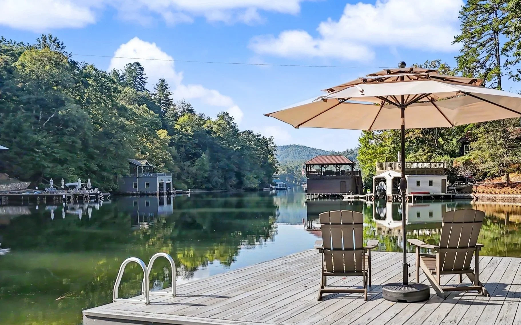 A Lake Burton vacation rental dock scene with a wooden deck, two Adirondack chairs, and a large beige patio umbrella. The calm water reflects surrounding trees and lake docks, with green foliage, blue sky, and scattered clouds in the background.