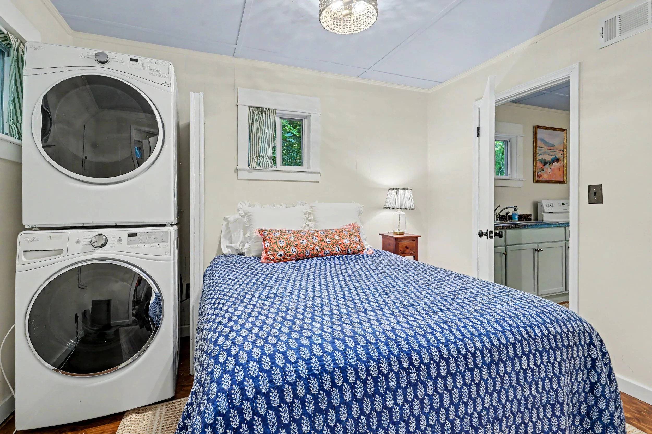 A small bedroom with a bed covered in a blue and white patterned quilt, a pillow with an orange and white pattern, a bedside table with a lamp, two small windows with striped curtains, and a wall-mounted painting of a landscape. A washer and dryer ar