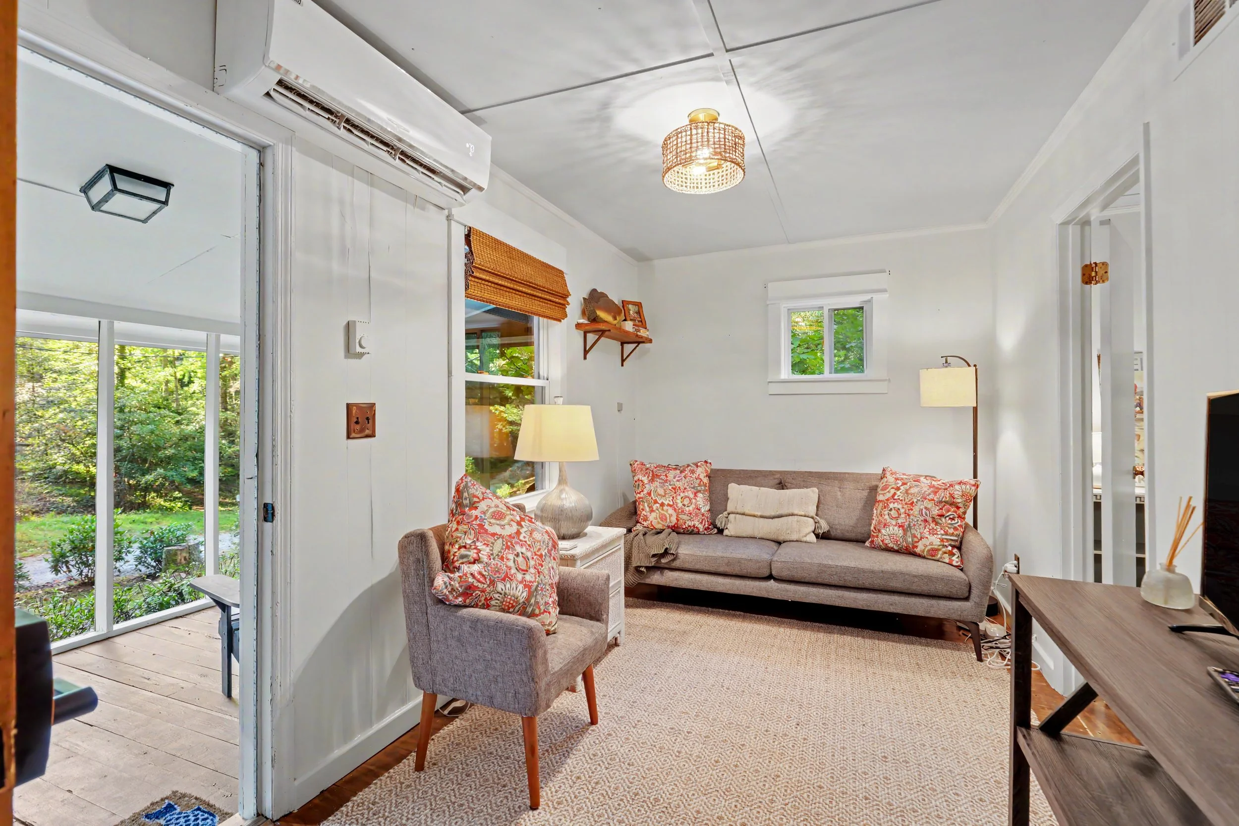 Living room with beige sofa, armchair, patterned pillows, wooden side table, lamp, wall shelf, windows with orange Roman shades, visible outdoor trees, and an air conditioning unit on the wall.