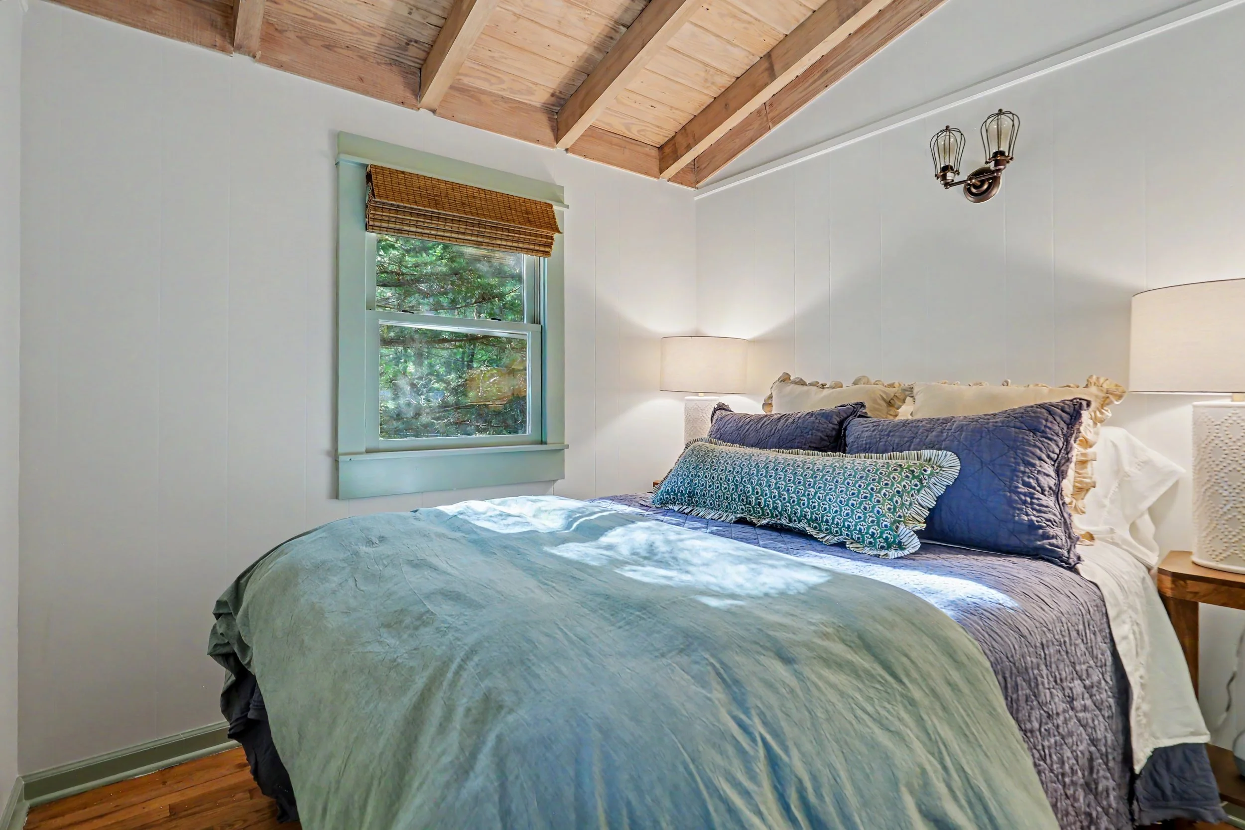 A cozy bedroom with a bed covered in a green quilt and decorated with patterned pillows. There is a large window with a view of trees, a bamboo shade, and a wooden ceiling with exposed beams. Two lamps flank the bed, and a small bedside table is part
