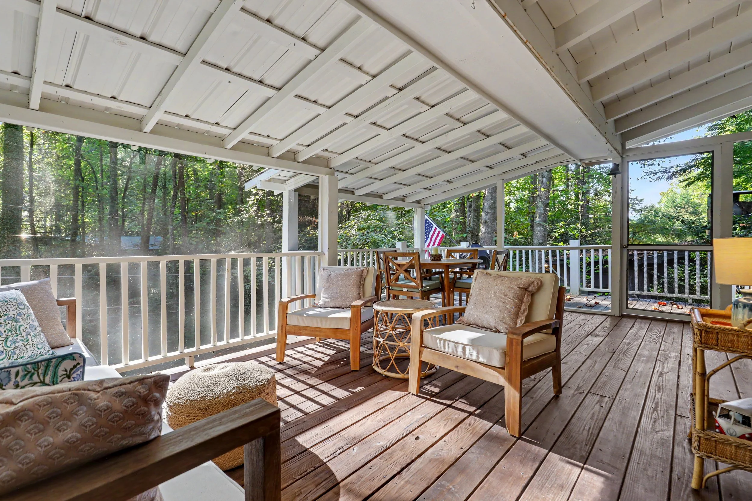 Covered outdoor porch with wooden flooring, white railing, seating with cushions, and an American flag, surrounded by green trees.