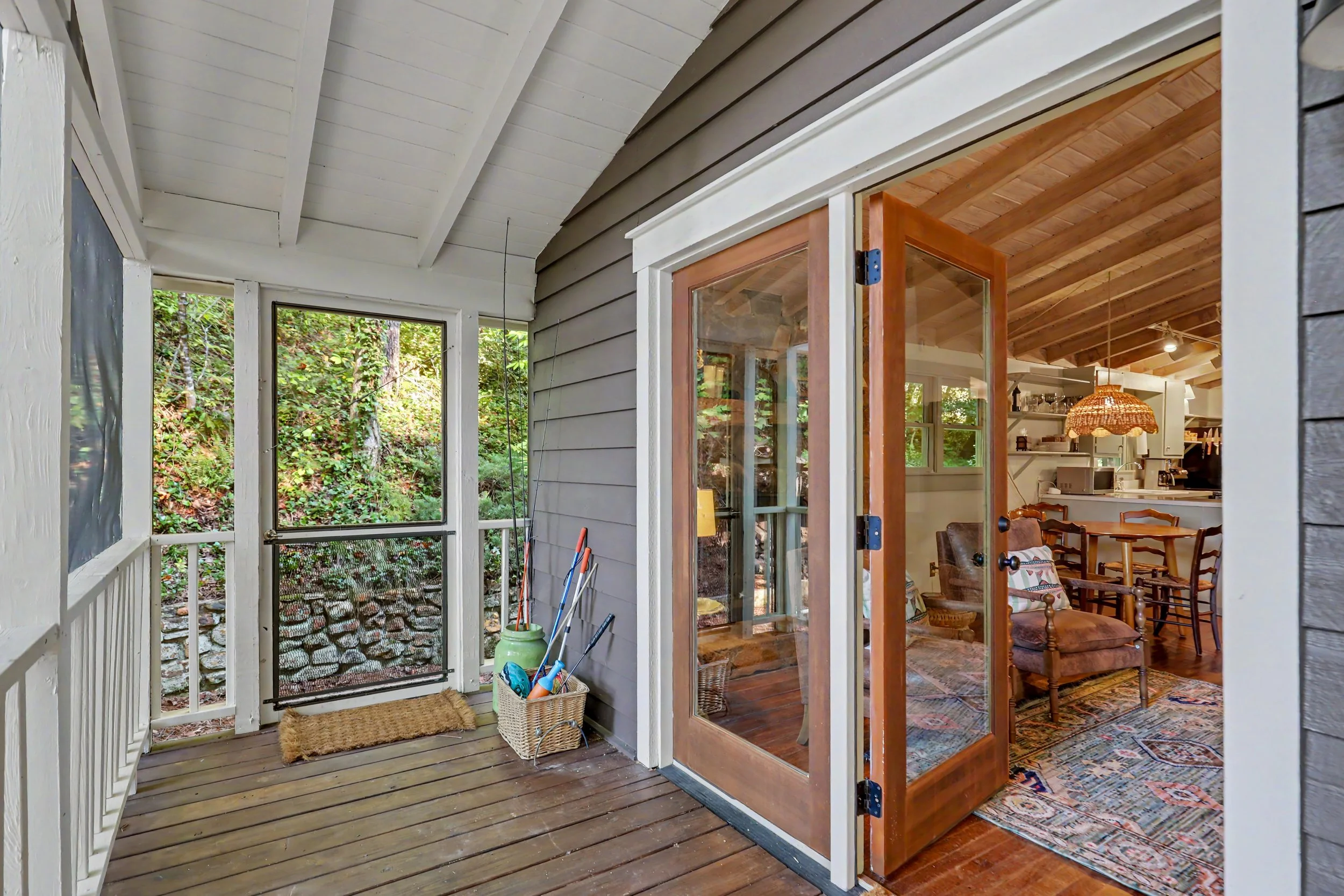 View of a screened-in porch area with a screened door, trailing plants outside, and an interior living room with wooden ceiling beams, a hanging lamp, and rustic furniture.