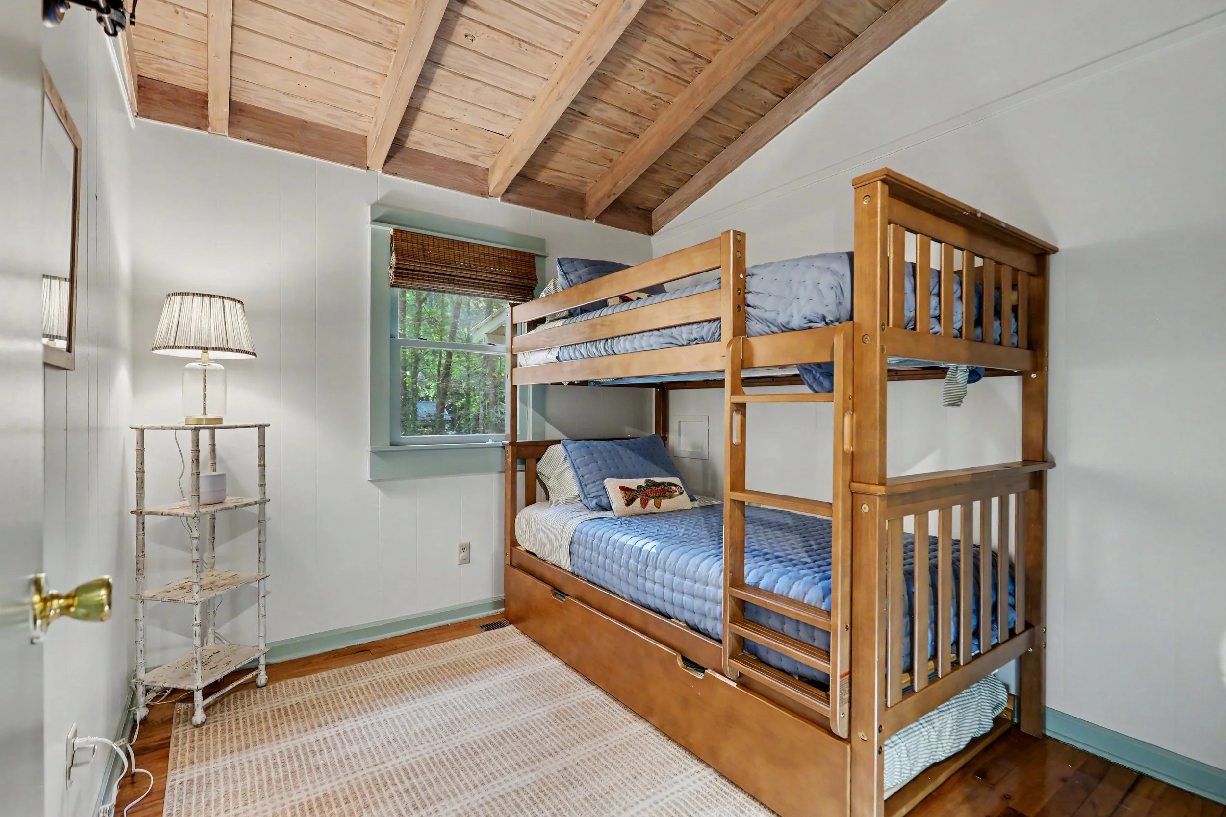 A small Lake Burton cabin bedroom with a wooden bunk bed, a window with a bamboo shade, a white lamp on a distressed white side table, and a beige rug on wood floor.