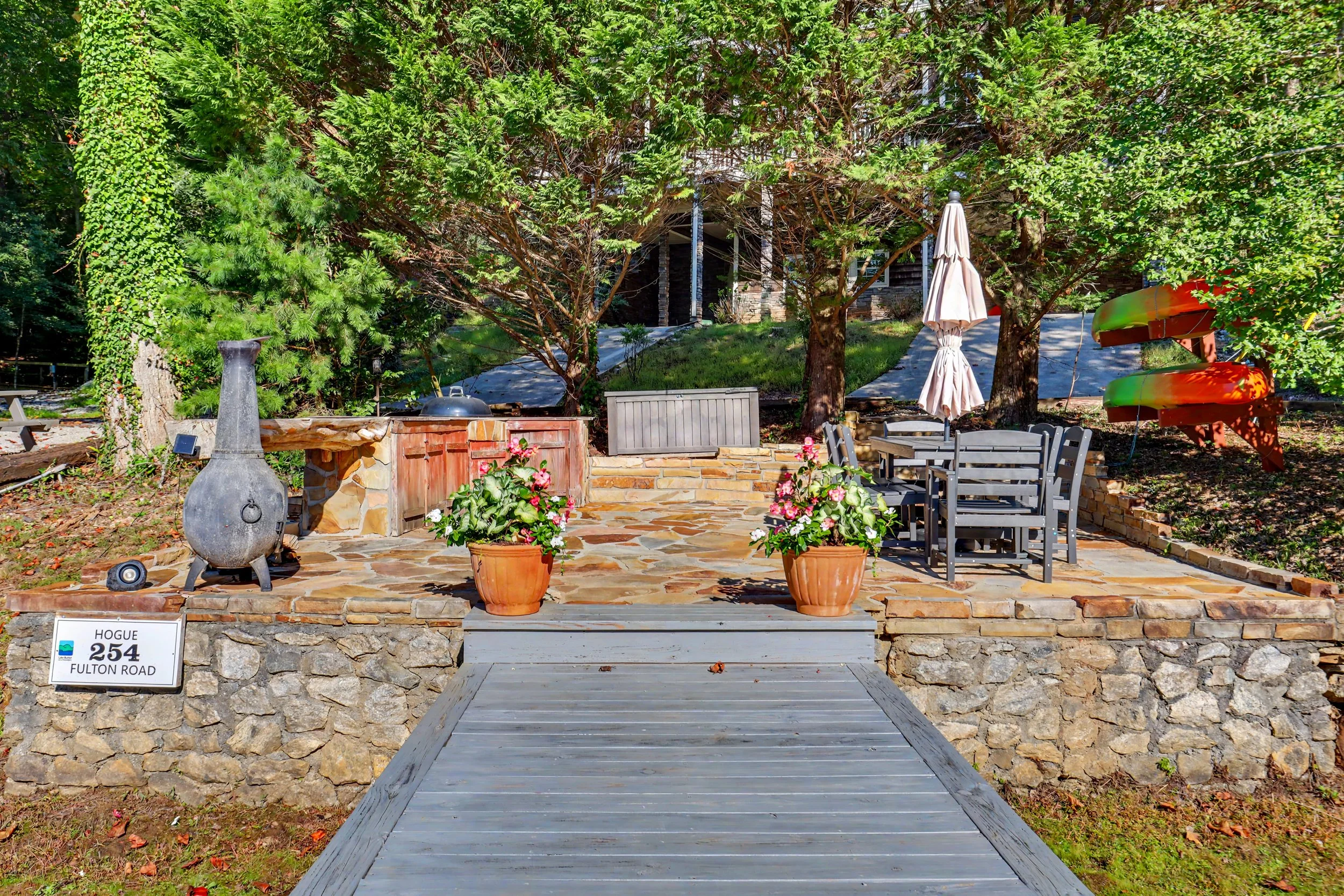 A lakefront patio area with a stone wall, wooden deck, potted plants, and outdoor furniture, surrounded by trees and greenery.