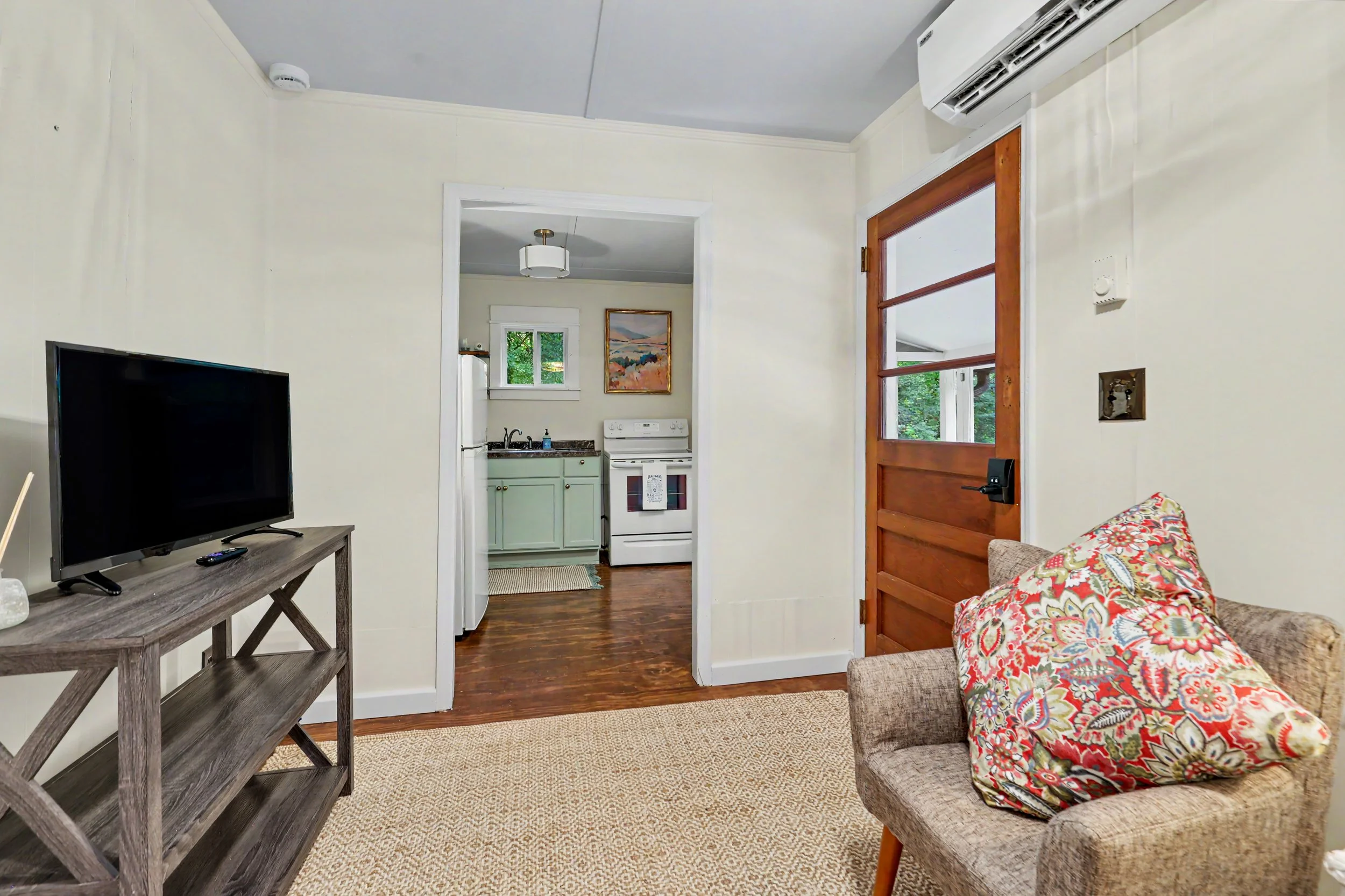 Living room with a TV on a wooden stand, a patterned chair, and a view into the kitchen with a sink, stove, and framed painting.