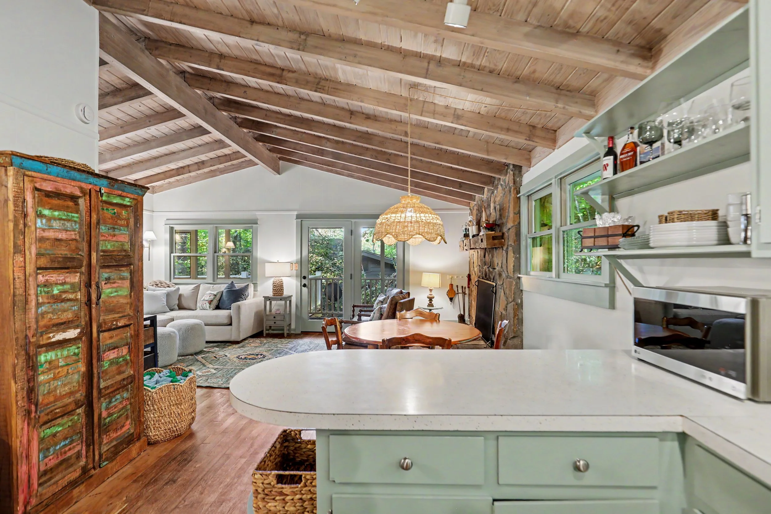 Interior view of a Lake Burton cabin rental living room with a vaulted wooden ceiling, a white sofa, a round dining table with wooden chairs, a stone accent wall, open shelves with glassware and bottles, and a rattan pendant light.