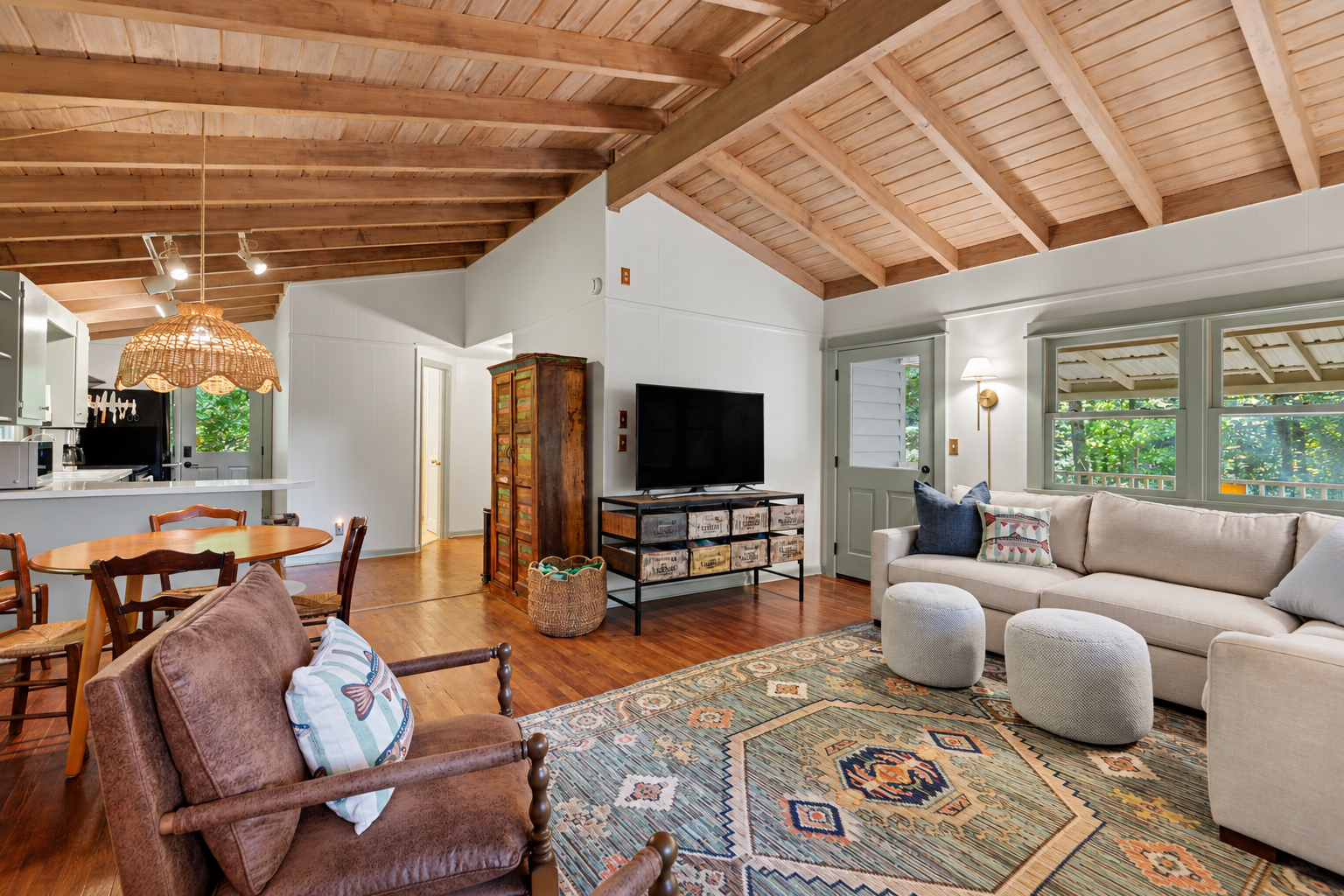 Living room with a beige sectional sofa, two round ottomans, a colorful patterned rug, a wall-mounted TV on a stand, a vintage wooden cabinet, and windows overlooking greenery. There is a dining table with chairs and a kitchen area with a hanging wic