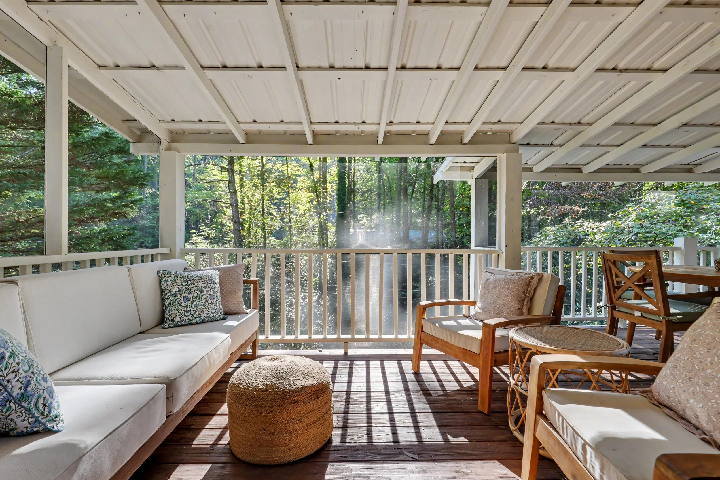 Screened-in porch with white ceiling, outdoor furniture including a white sofa, wooden chairs, and a woven ottoman, surrounded by North Georgia mountain trees and greenery.