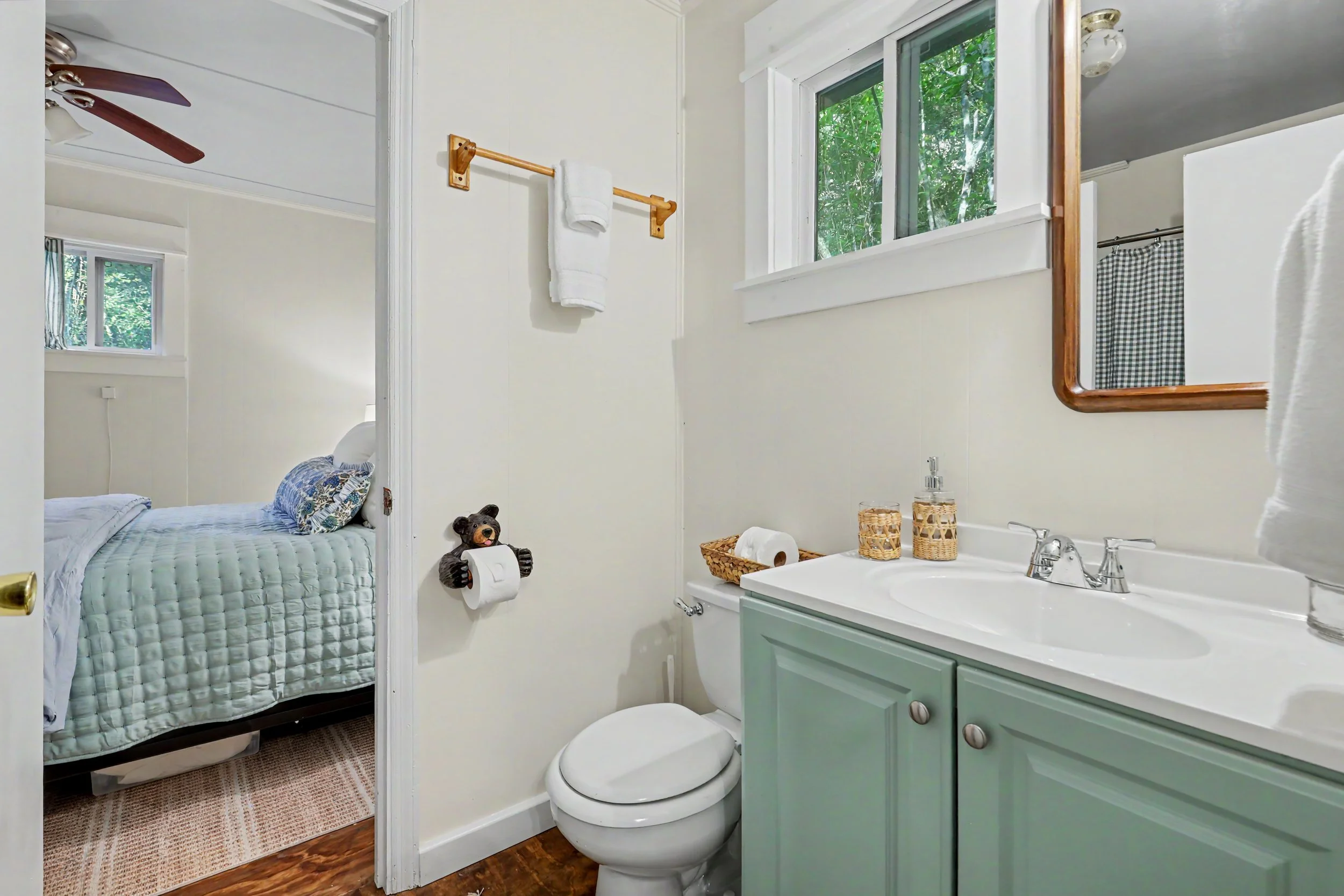 A small bathroom in Lake Burton vacation rental cottage with a white sink and green cabinet, a mirror, and a window with greenery outside. The bathroom has a toilet, wicker baskets, and a towel on a wooden towel rack.