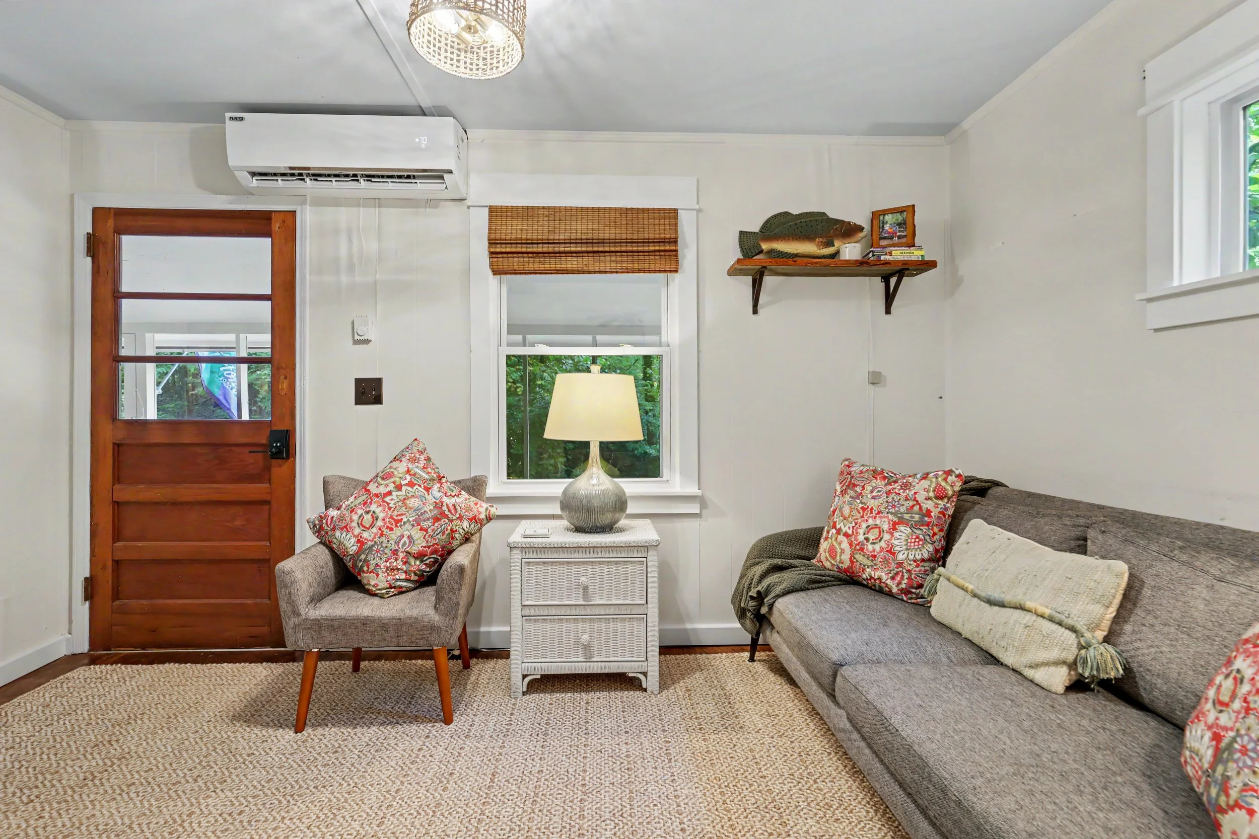 A Lake Burton cabin rental living room with a red wooden door, sofa with floral pillows, a chair with a similar floral pillow, a white wicker side table with a lamp and a window.