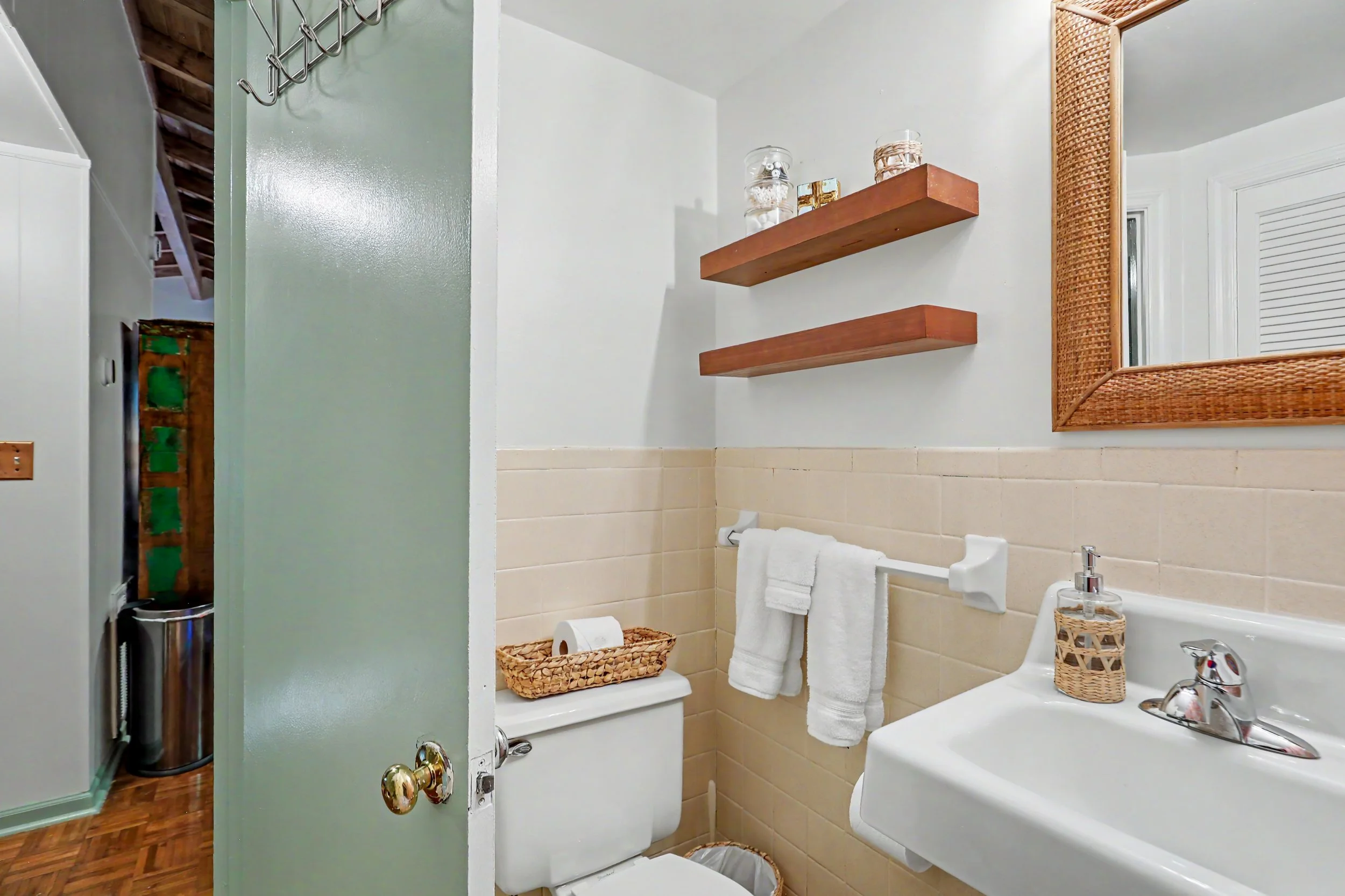 A small bathroom with beige tiled walls and a white toilet, above it a wicker basket, a wooden shelf with decorative jars, a large mirror with a wicker frame, a white sink, and a bottle of hand soap.