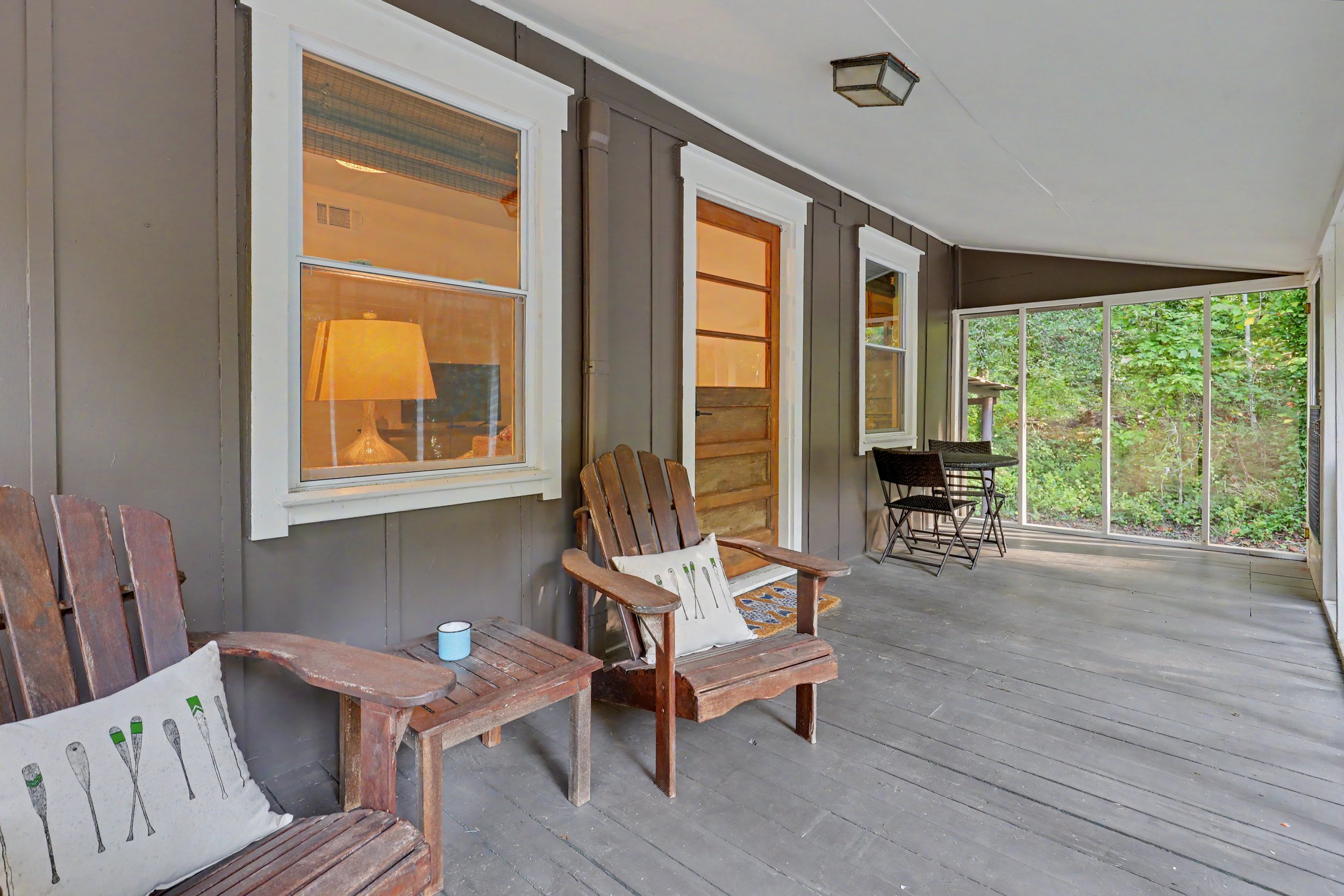 A covered porch with wooden chairs, pillows, a lamp inside, and a view of greenery outside.
