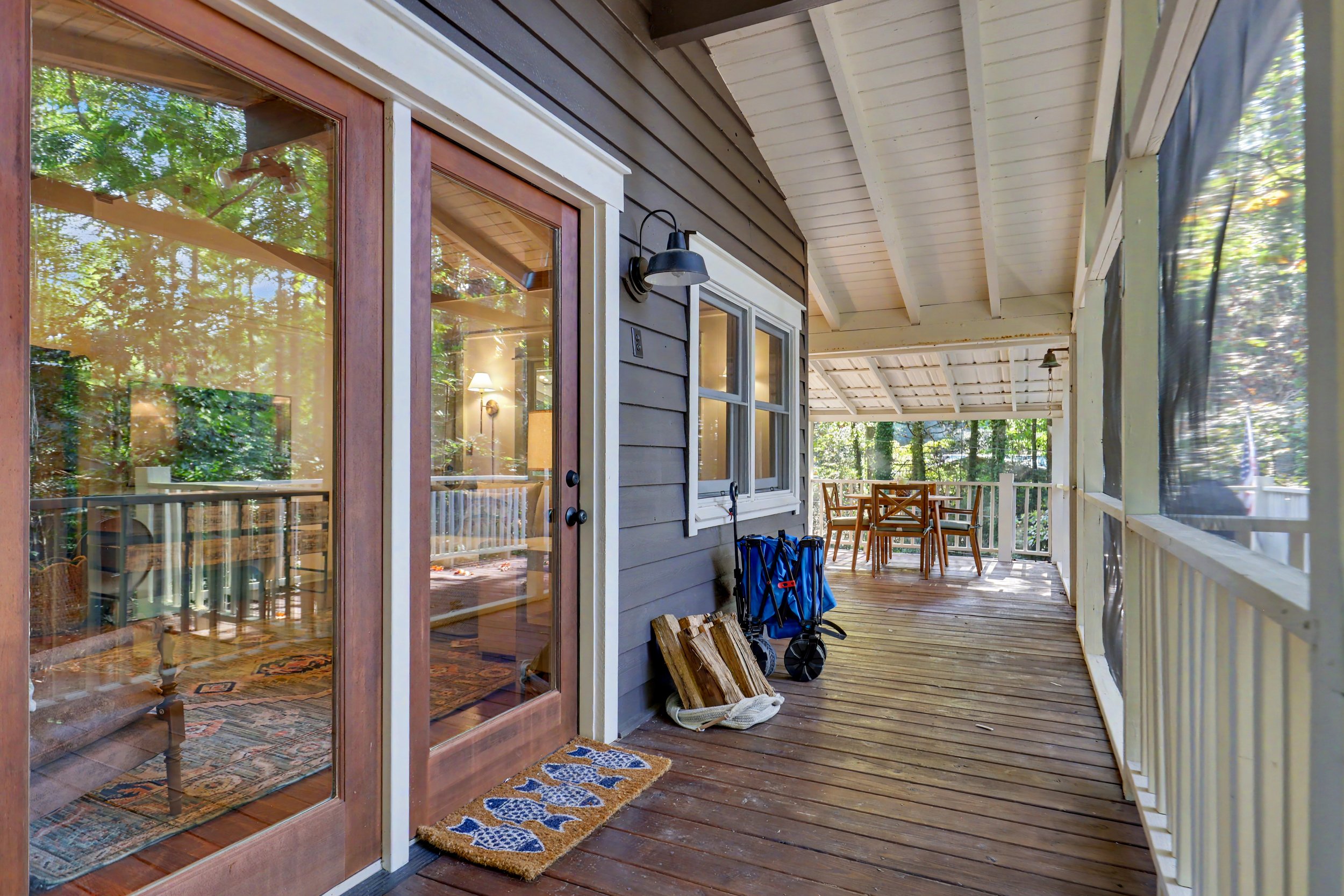View of a screened porch with a wooden deck, sliding glass door leading into a home interior, outdoor furniture, and green trees in the background.