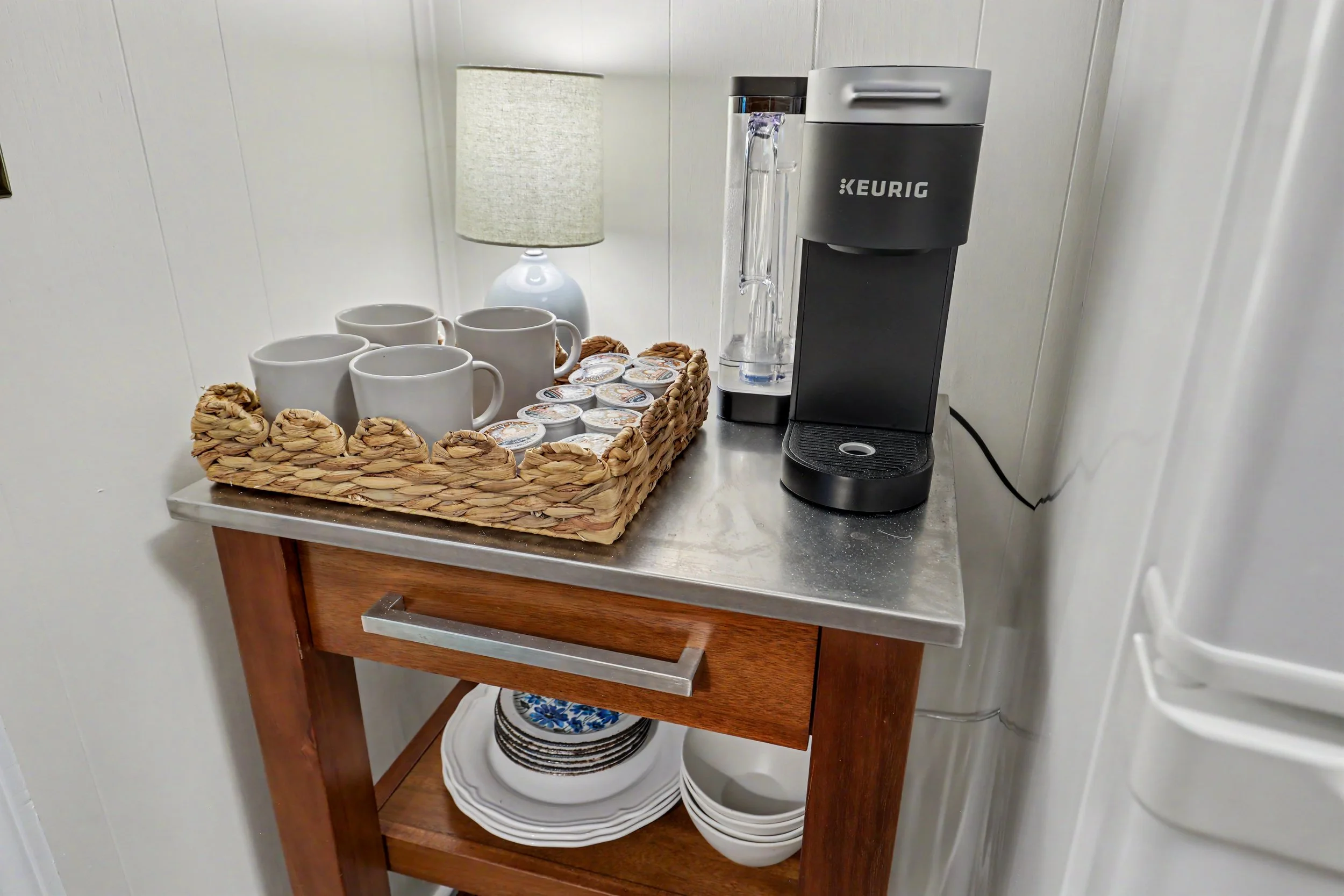 A kitchen countertop with a basket of coffee mugs, small containers of creamers, a small lamp, and a Keurig coffee machine. The shelf below holds white bowls and plates.