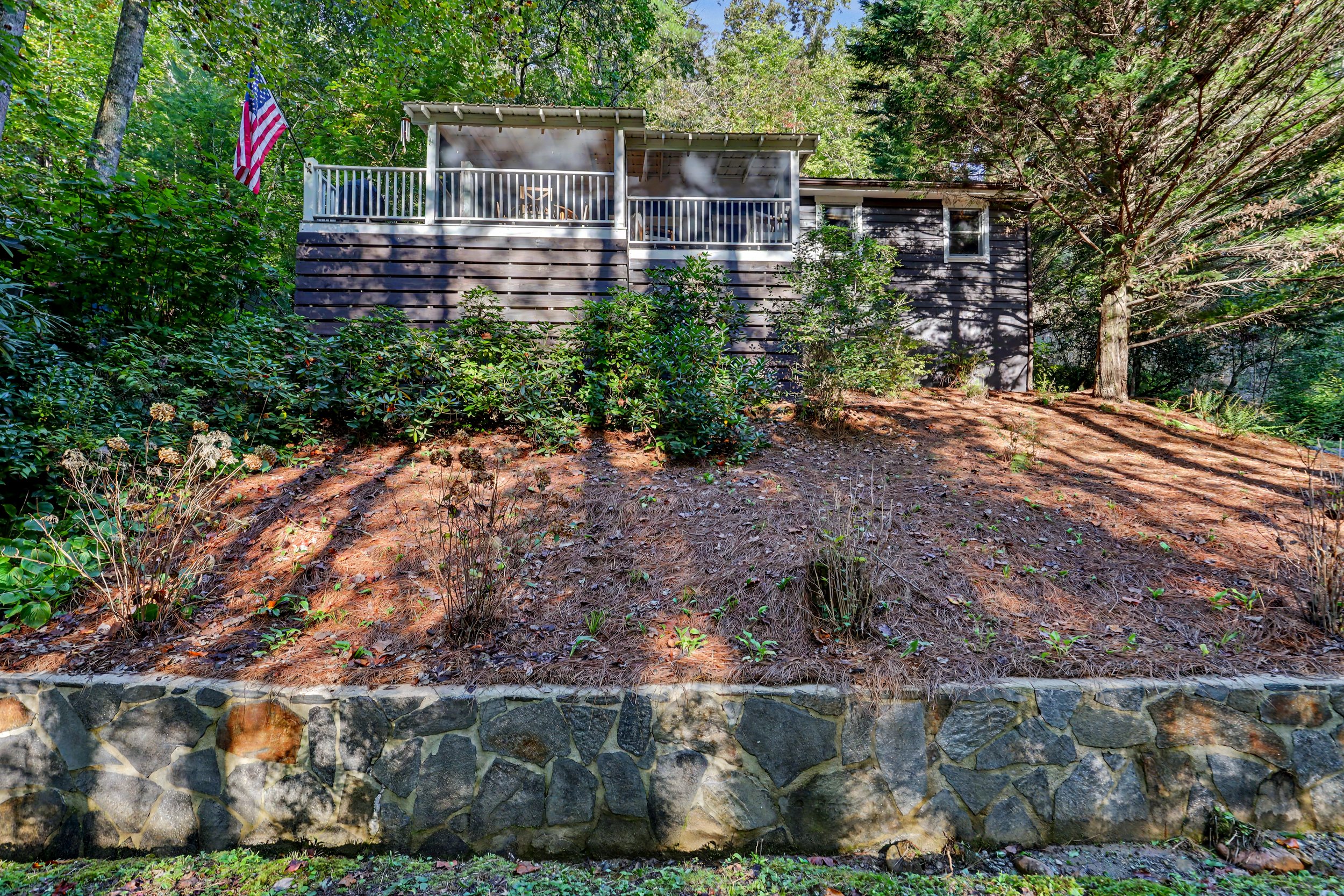 A black wooden Lake Burton mountain house with a covered porch, surrounded by green trees and bushes, with an American flag hanging on the left side.