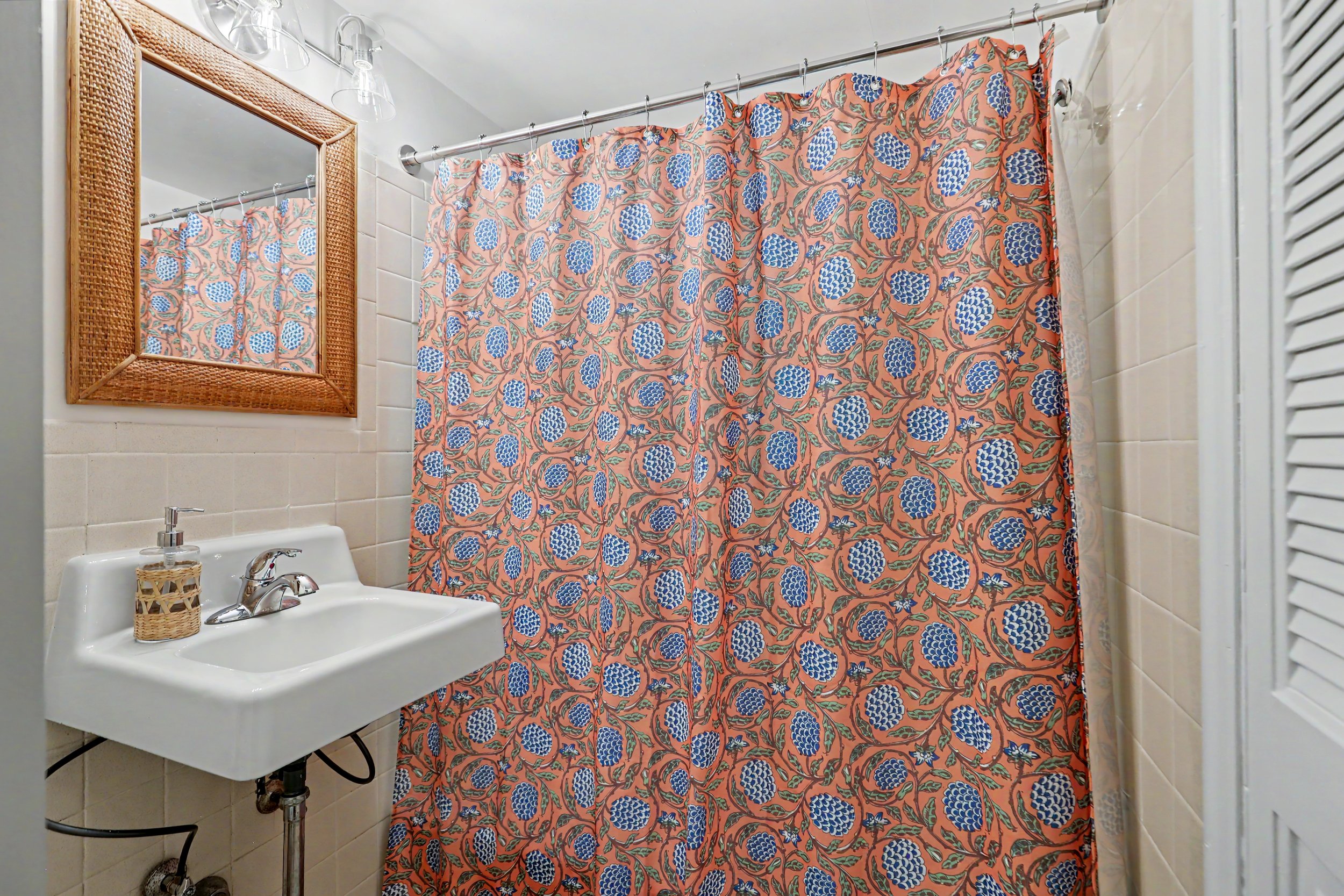 Bathroom with a woven-framed mirror, a white sink with a soap dispenser, and a floral shower curtain with blue and orange pattern.
