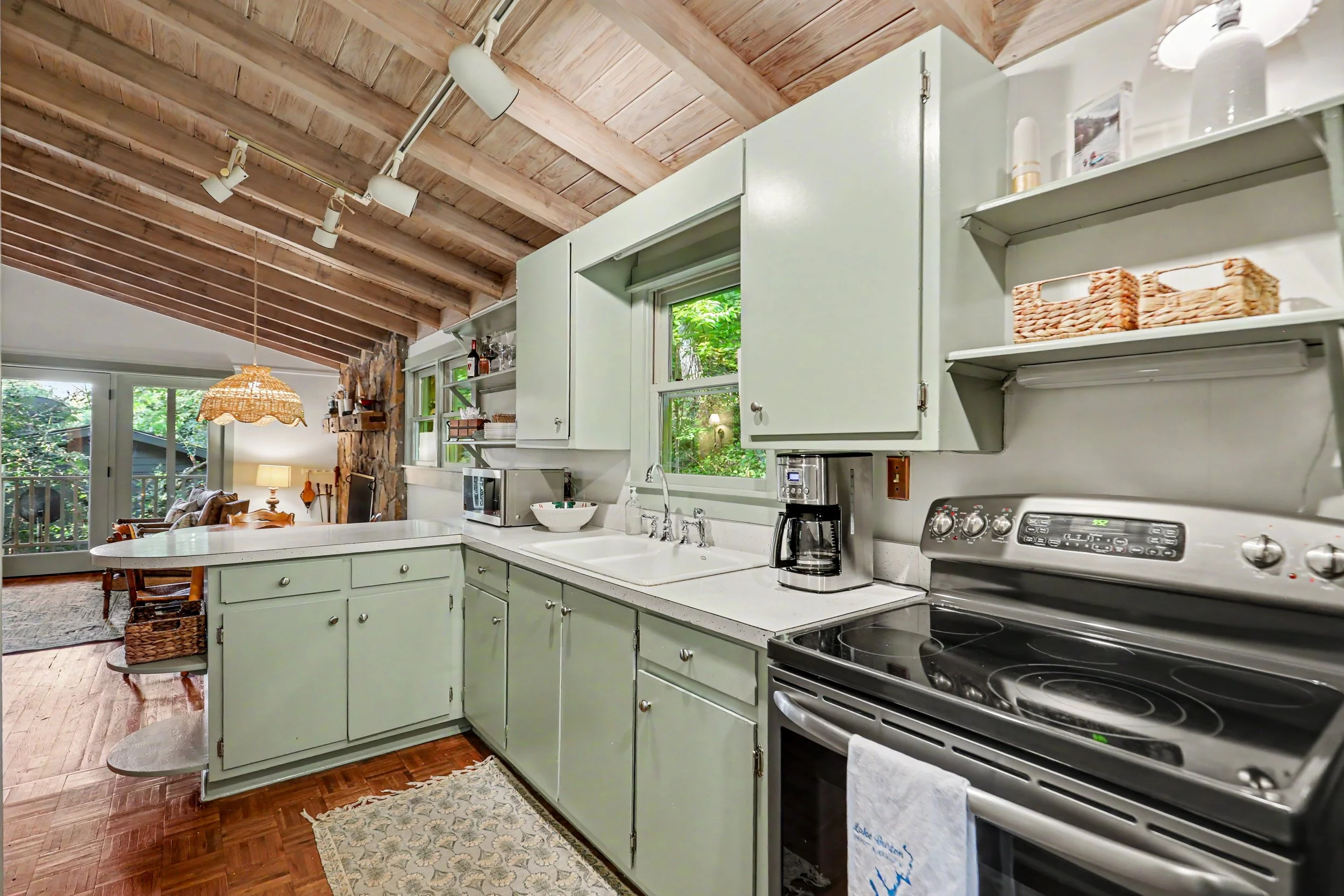 Kitchen with green cabinets, white countertops, a stove, coffee maker, microwave, and a window showing outside greenery. There's a living area with a lamp and dining table in the background, and a wooden ceiling.