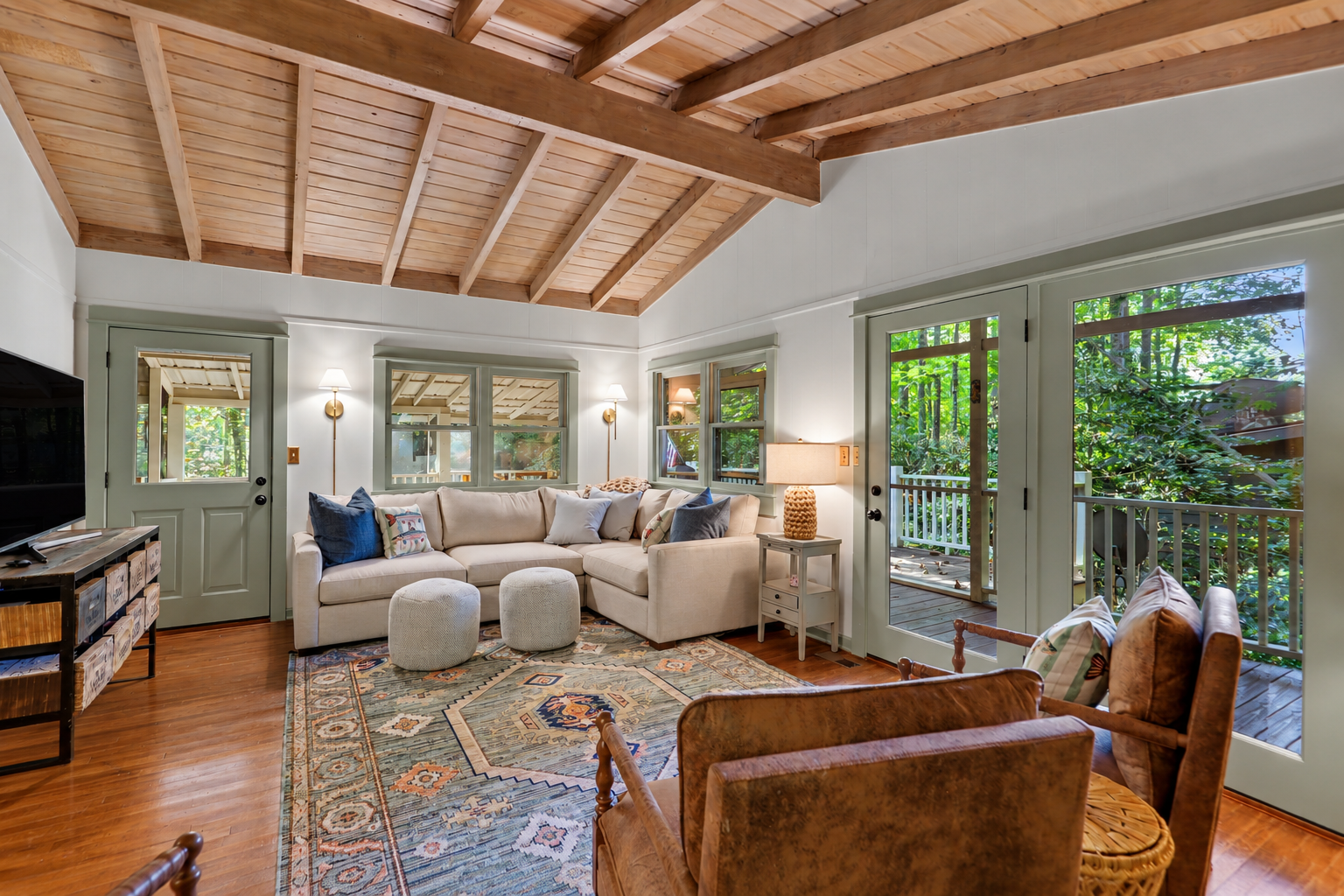 Cozy living room with beige sectional sofa, two small white poufs, patterned area rug, wooden floor, and a wood-paneled vaulted ceiling. Green framed glass door and windows reveal a lush outdoor scene. Side table with lamp and wall-mounted lights alo