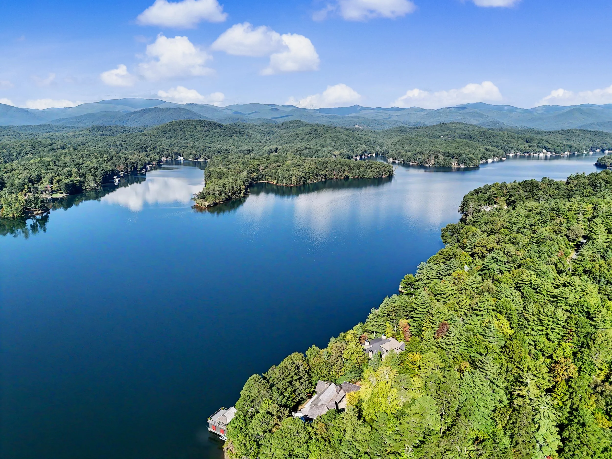 Aerial view of a Lake Burton with green, forested islands and surrounding lush trees, with mountains in the background and a partly cloudy sky.