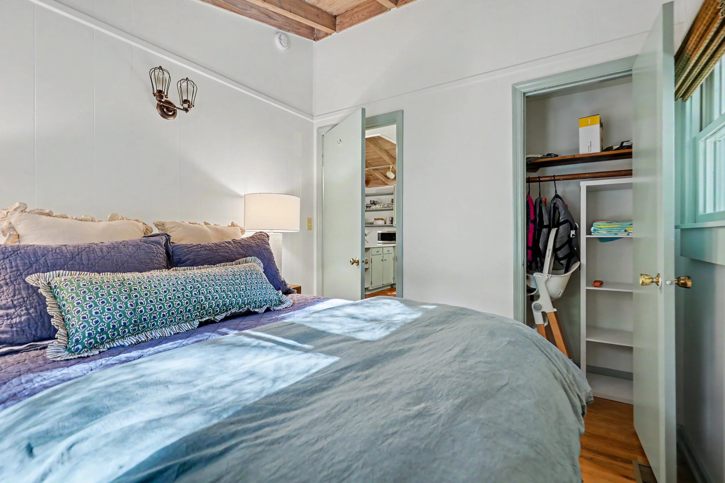 A bedroom with a bed featuring multiple pillows and a quilt, an open closet with clothing and a box, a small white chair, a lamp, and a view into an adjacent room with kitchen cabinets and a ceiling with wooden beams.
