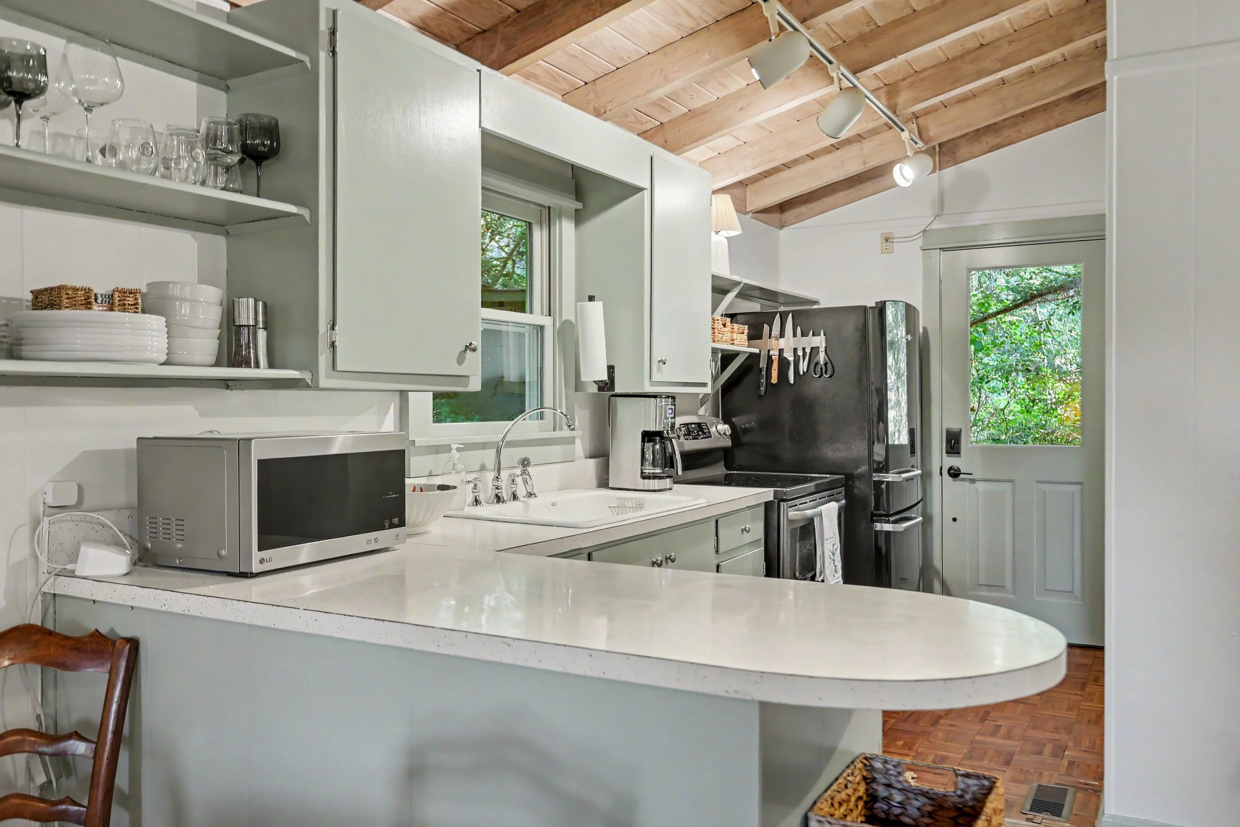 A kitchen with white cabinets, a white countertop, and a black refrigerator. There are open shelves with glasses and bowls, a microwave, a coffee maker, and a sink. A door and windows let in natural light, showing greenery outside.